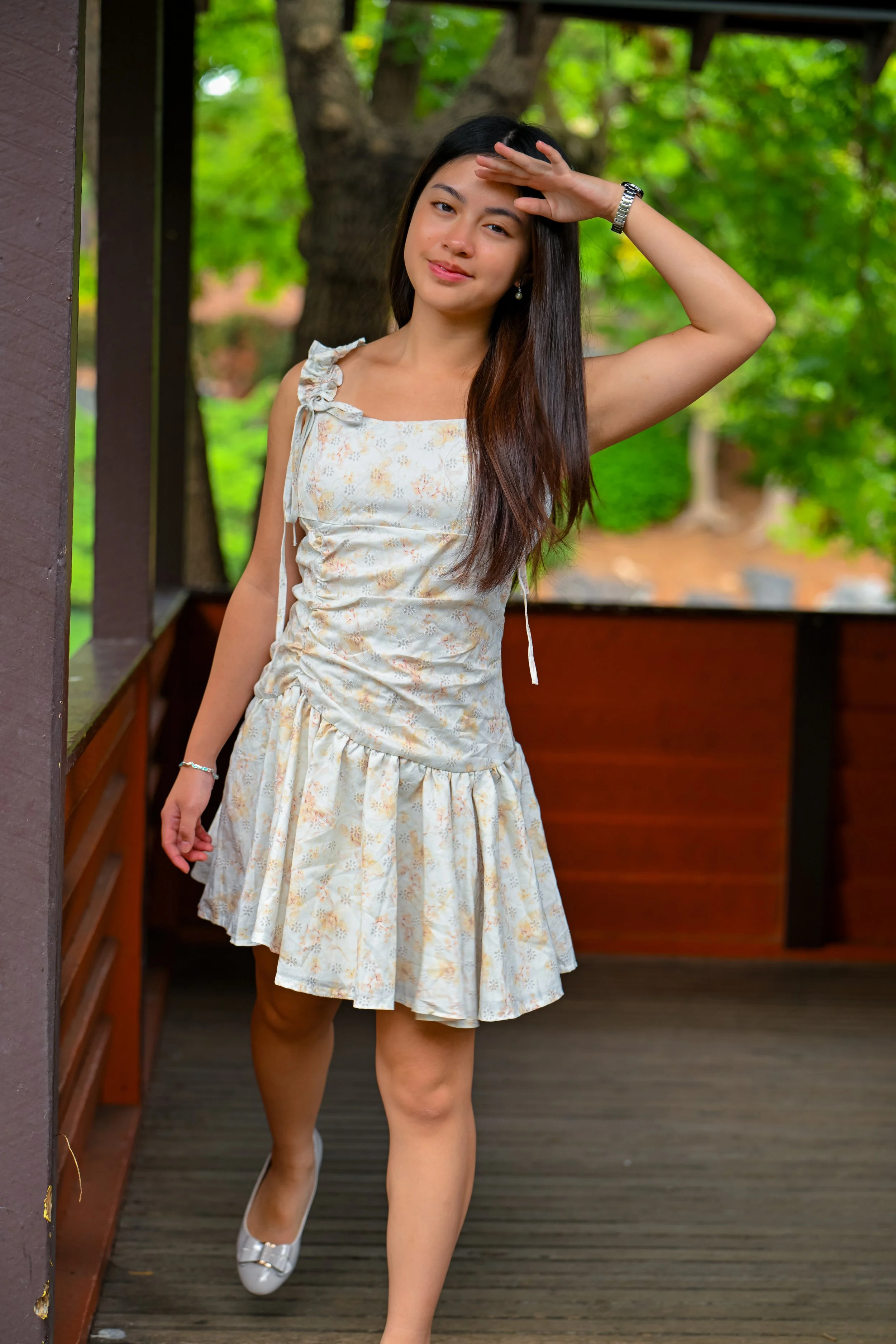 A young woman with long dark hair standing on a wooden balcony or porch, wearing a white floral dress and silver shoes, holding her hand above her eyes to shield them from the sun, with a background of green trees and a wooden railing.