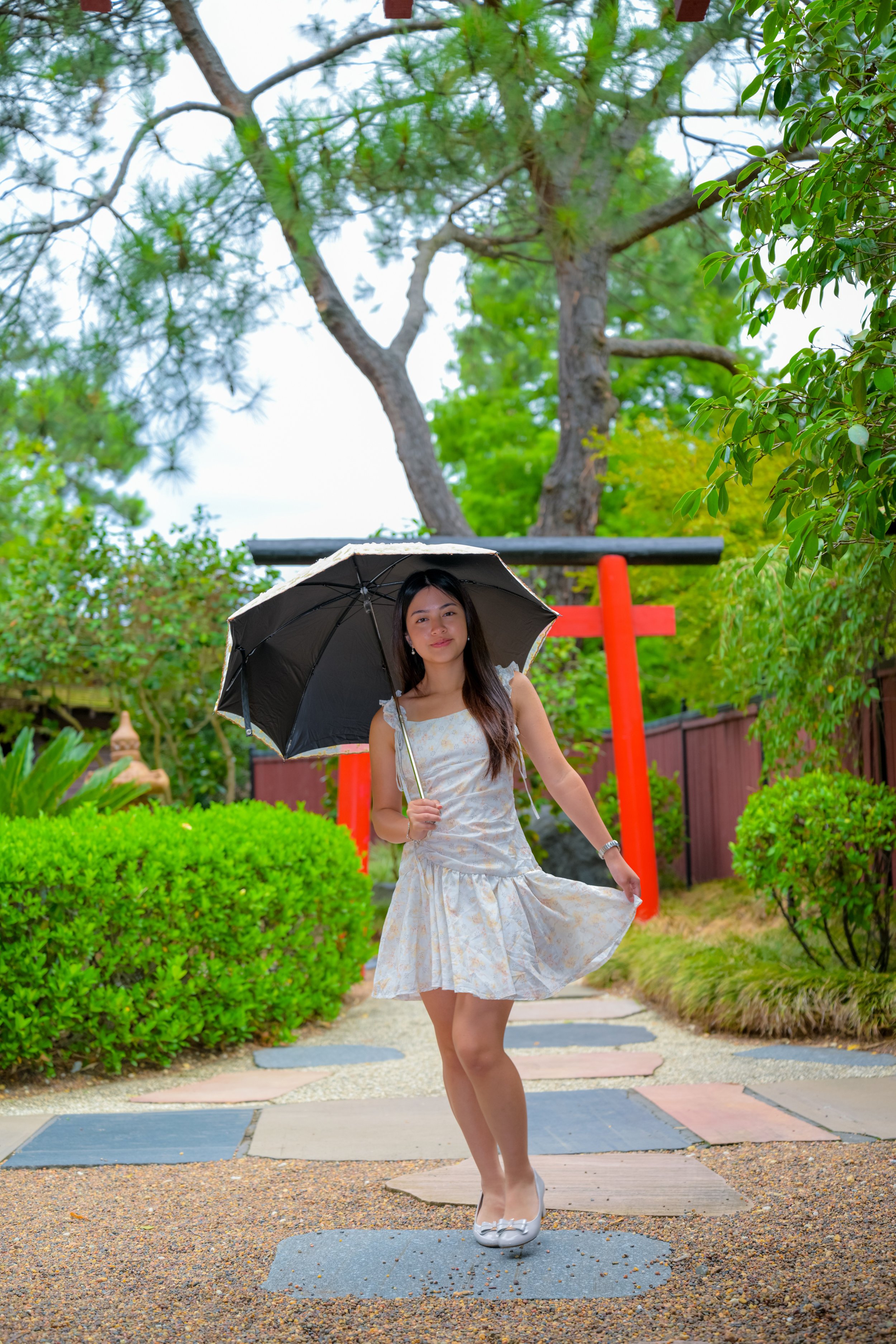 A young woman in a light-colored dress standing on a pathway in a garden, holding an umbrella, with a red torii gate and green trees in the background.