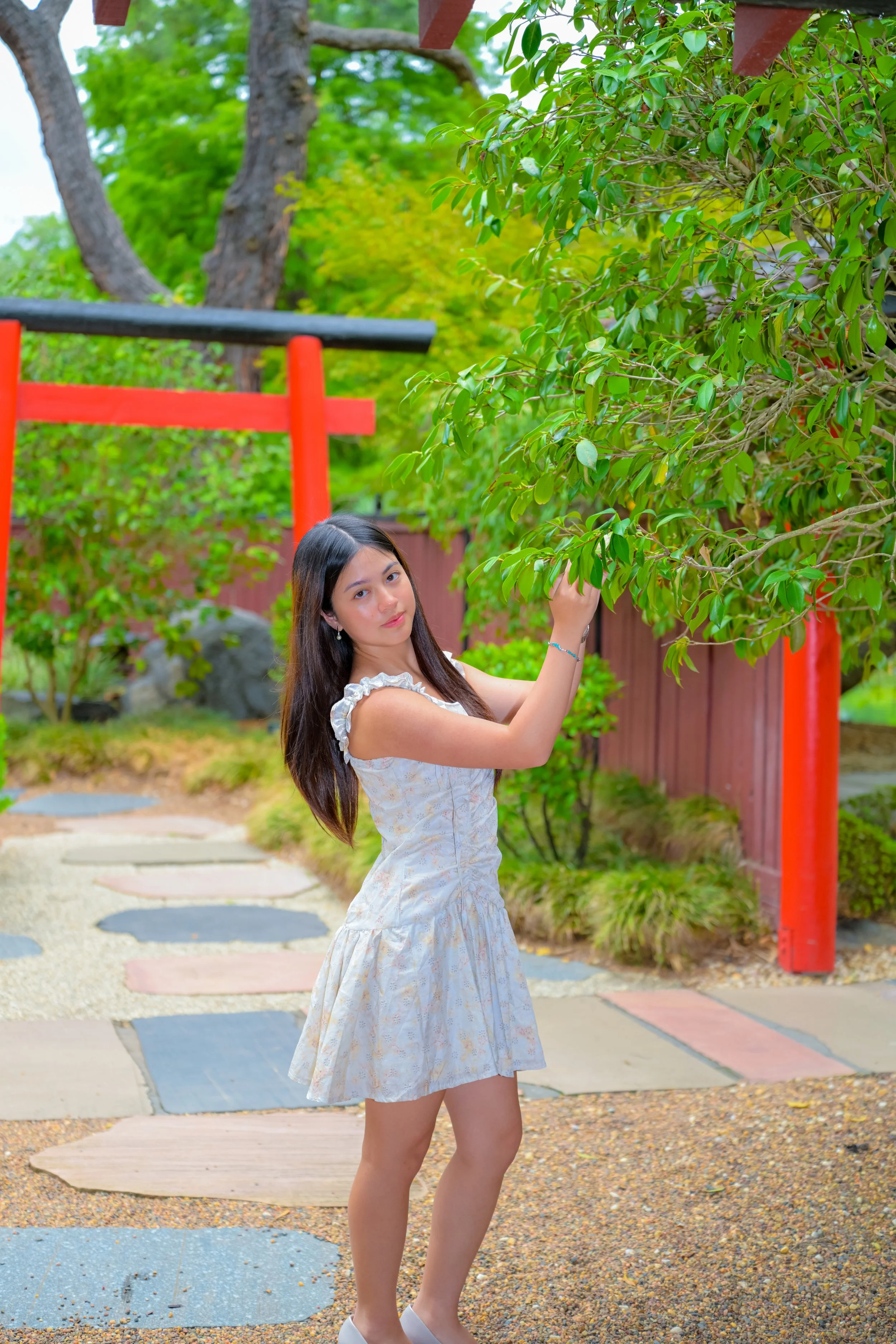 A young woman in a light-colored dress standing outdoors near a tree, with a Japanese torii gate and a wooden fence in the background, surrounded by greenery.