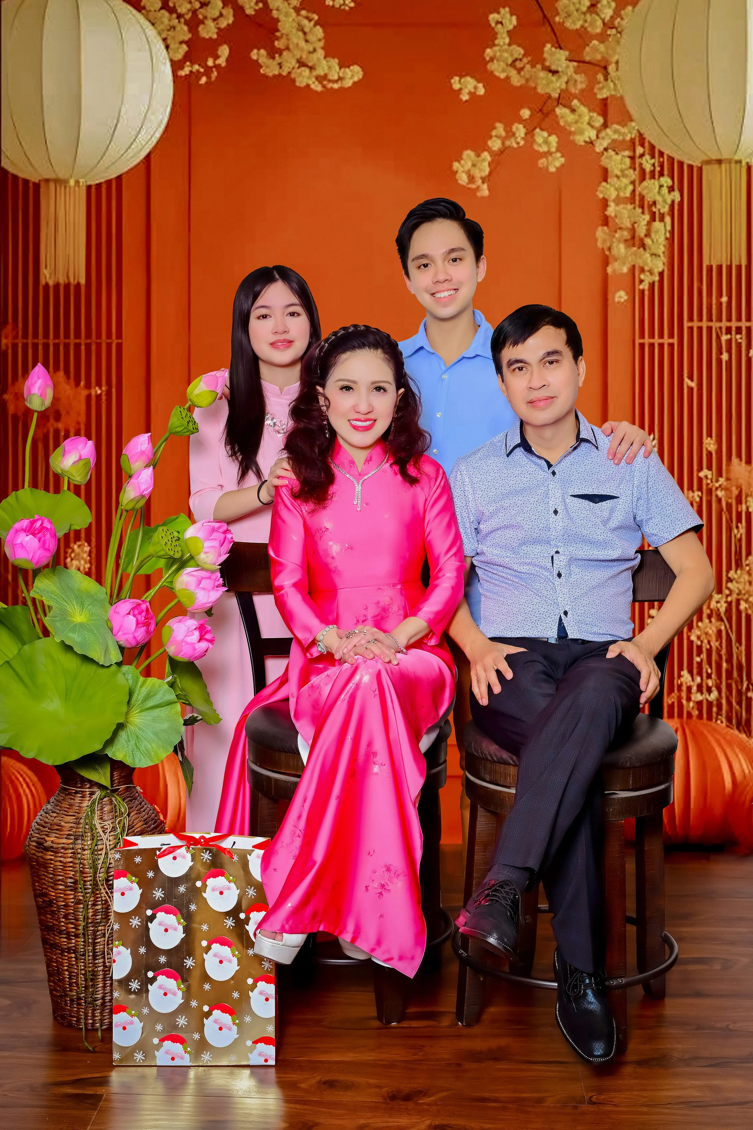 A family of five posing for a holiday photo with a red background, white flowers, lanterns, a large potted plant with pink flowers, and a gift bag with Santa and snowmen designs.