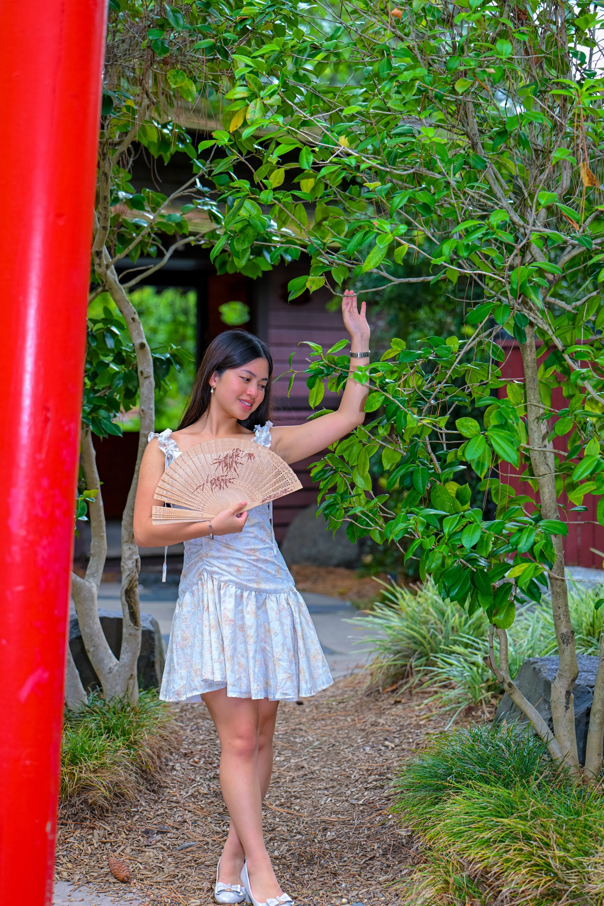 A woman in a white patterned dress holding a folding fan and touching a leaf on a tree in a garden.