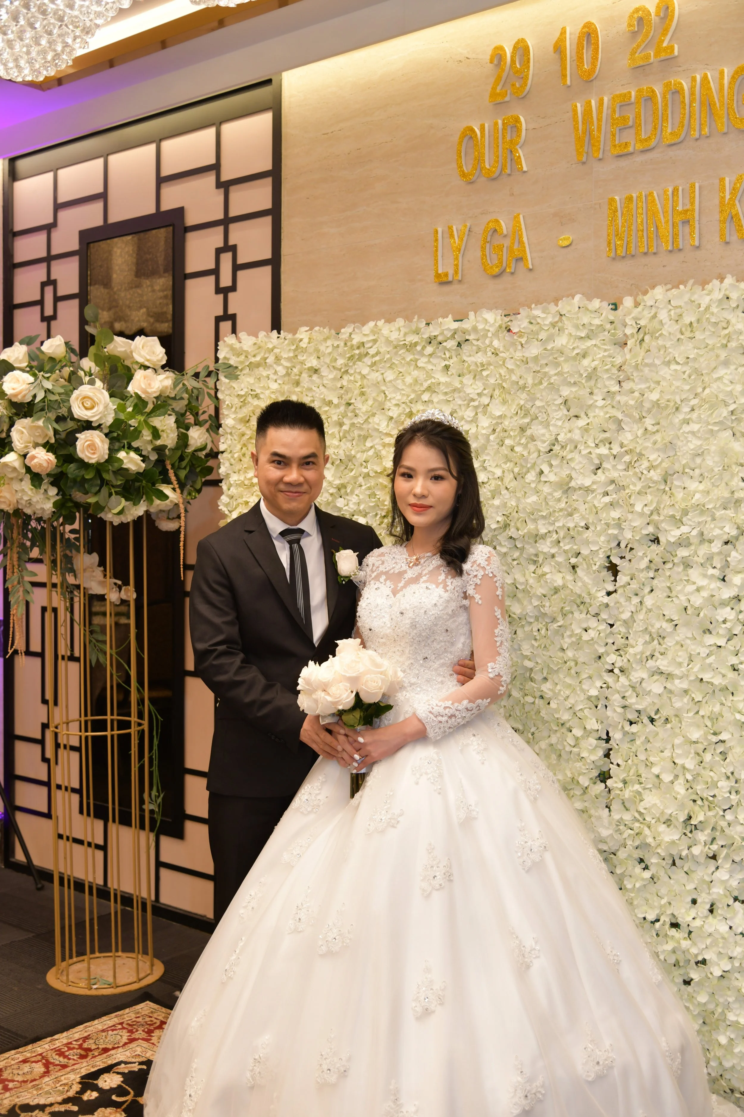 Wedding couple posing in front of a floral backdrop at their wedding reception with gold text on the wall indicating the date and location of the wedding.