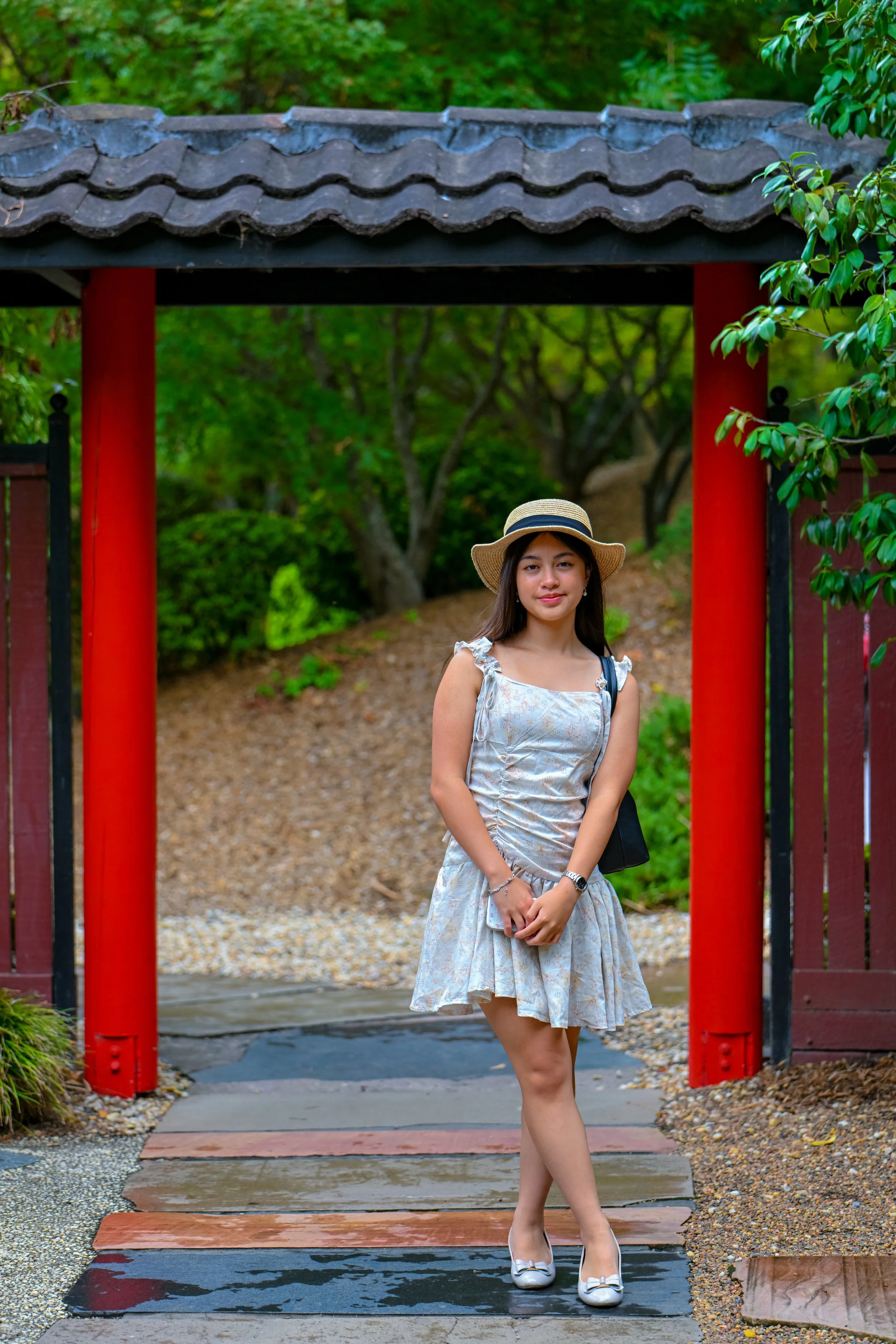 A young woman wearing a floral dress and a straw hat standing on a pathway under a traditional Asian-style gate, surrounded by green foliage.