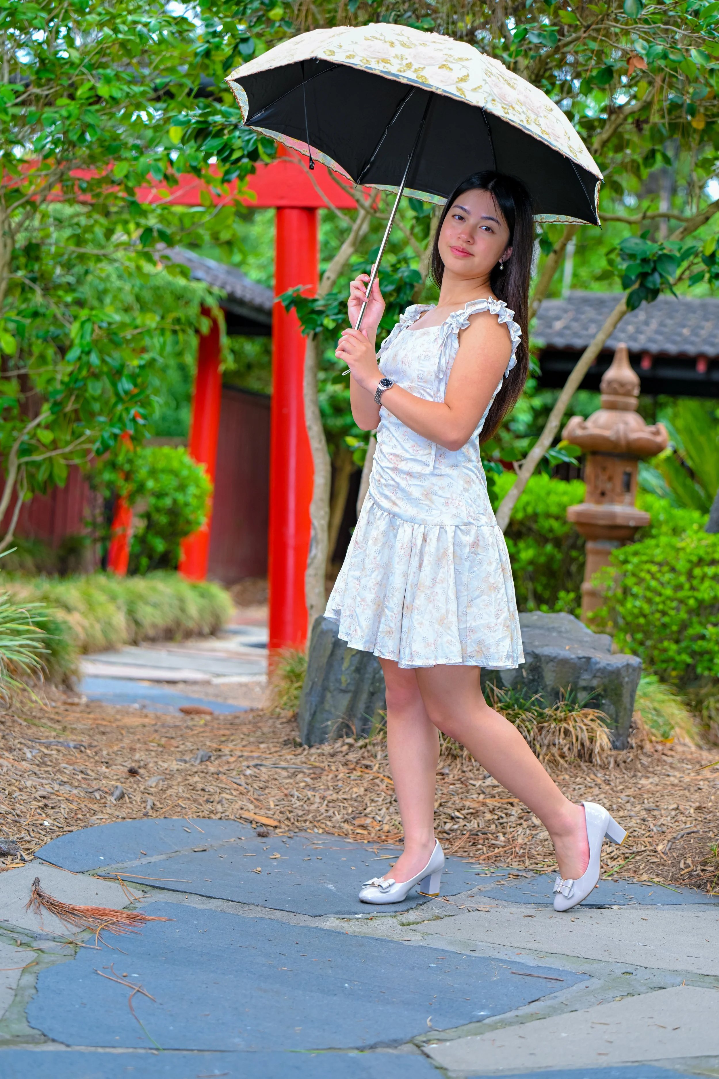 A young woman in a white floral dress and heels holding an umbrella, standing outdoors in a lush garden with traditional Japanese ornaments and red torii gates.