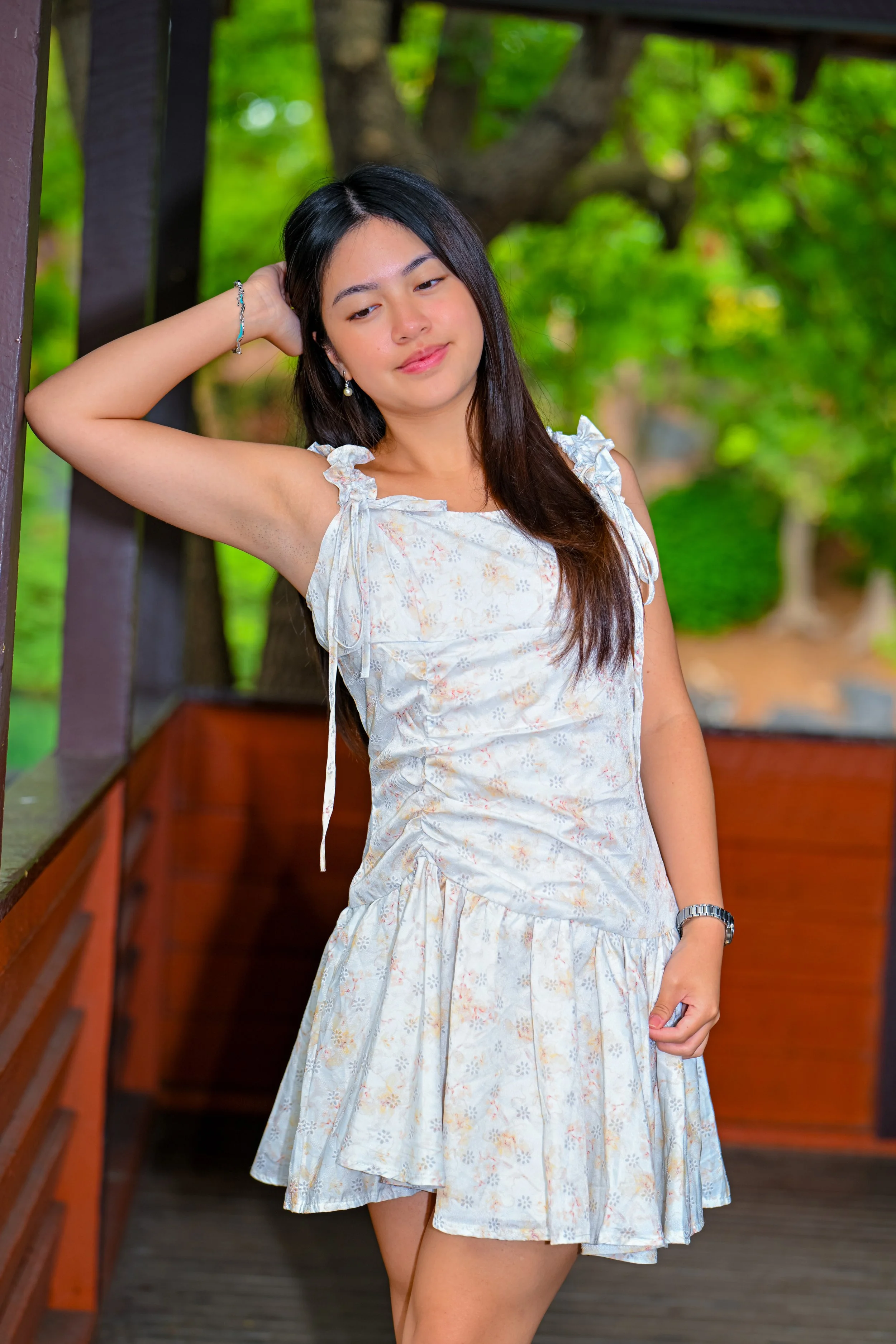 A young woman with long dark hair, wearing a light-colored floral dress, poses outdoors on a wooden deck with her hand behind her head, amidst green trees.