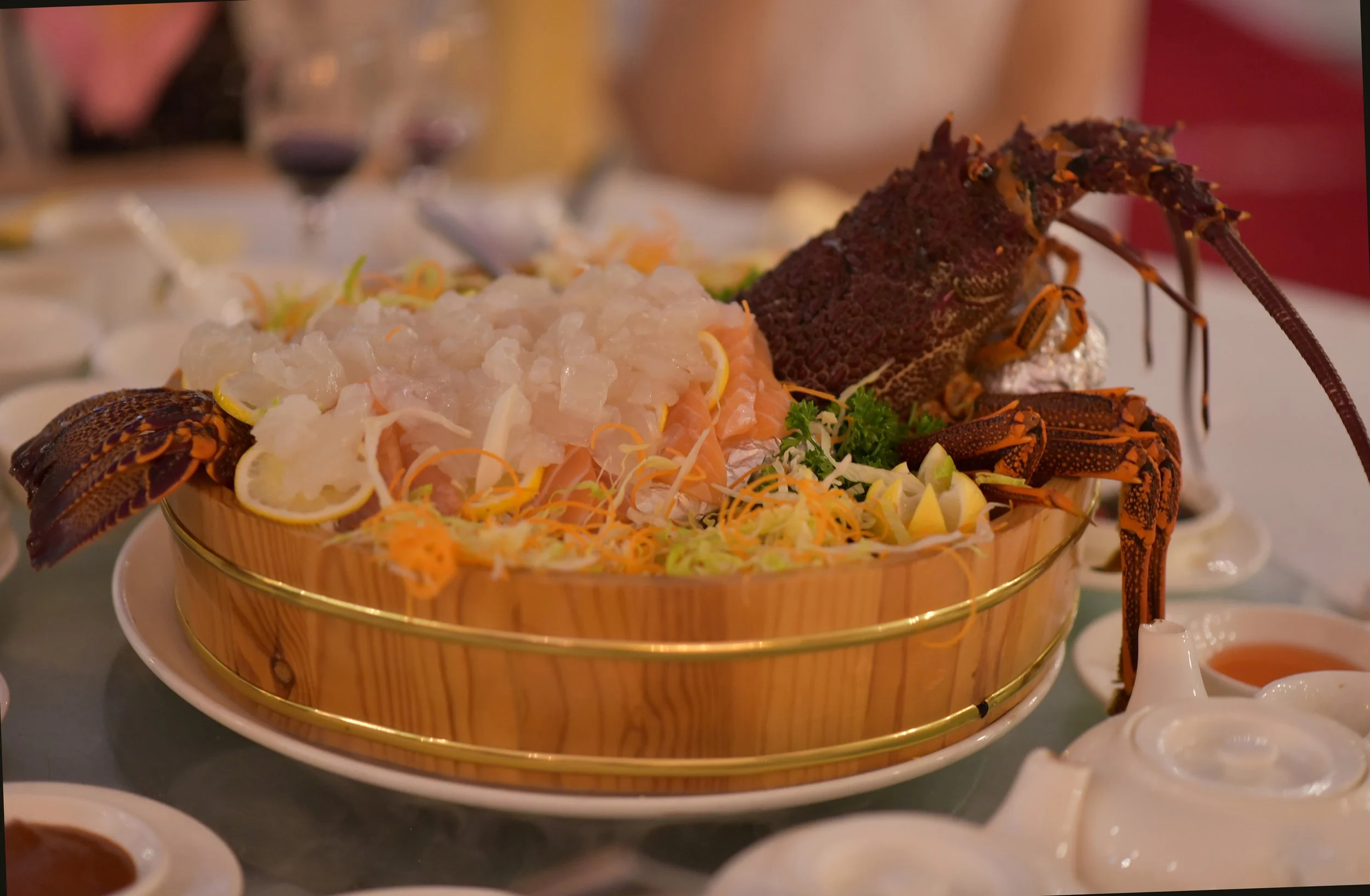 Lobster and sashimi on a platter with lemon slices and shredded vegetables, served in a wooden basket at a dinner setting.