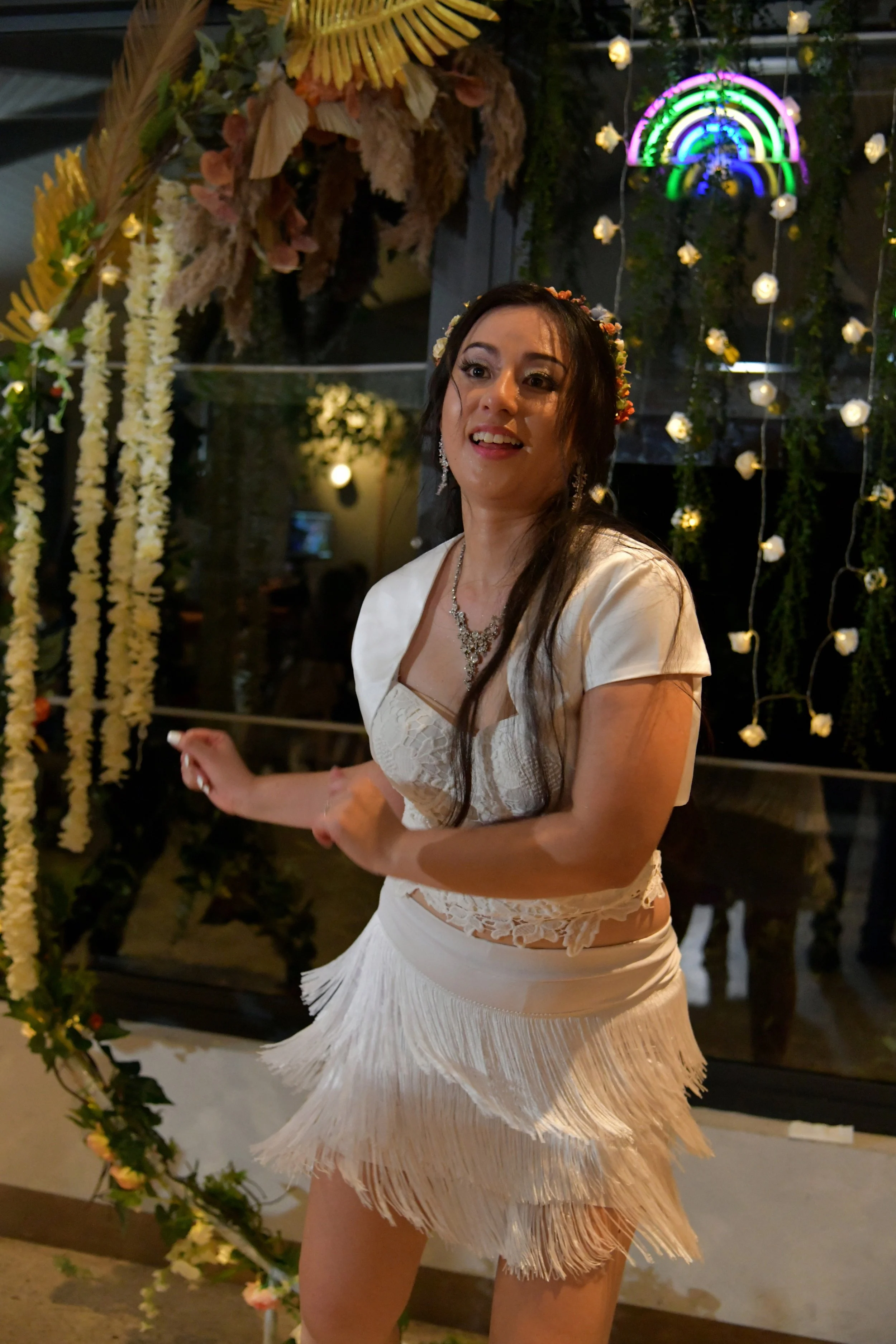 A woman in a white dress dancing in front of a floral and light decorated background at a celebration event.