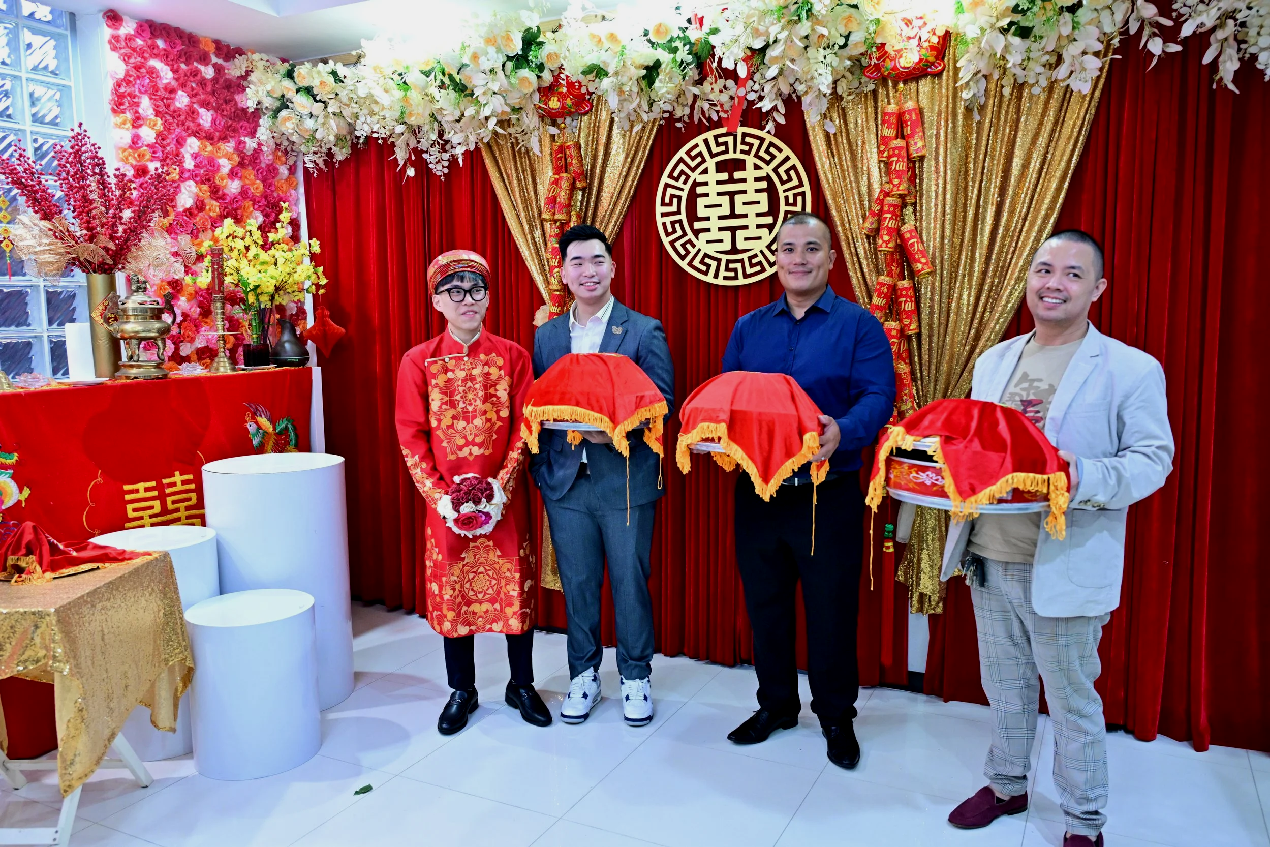Four men standing in front of a red and gold Chinese wedding backdrop, holding red covered objects. One man is dressed in traditional red attire with embroidered gold patterns and a head covering, while the others are in modern clothing. The backdrop features floral decorations, gold curtains, and a large Chinese character symbol in the center.