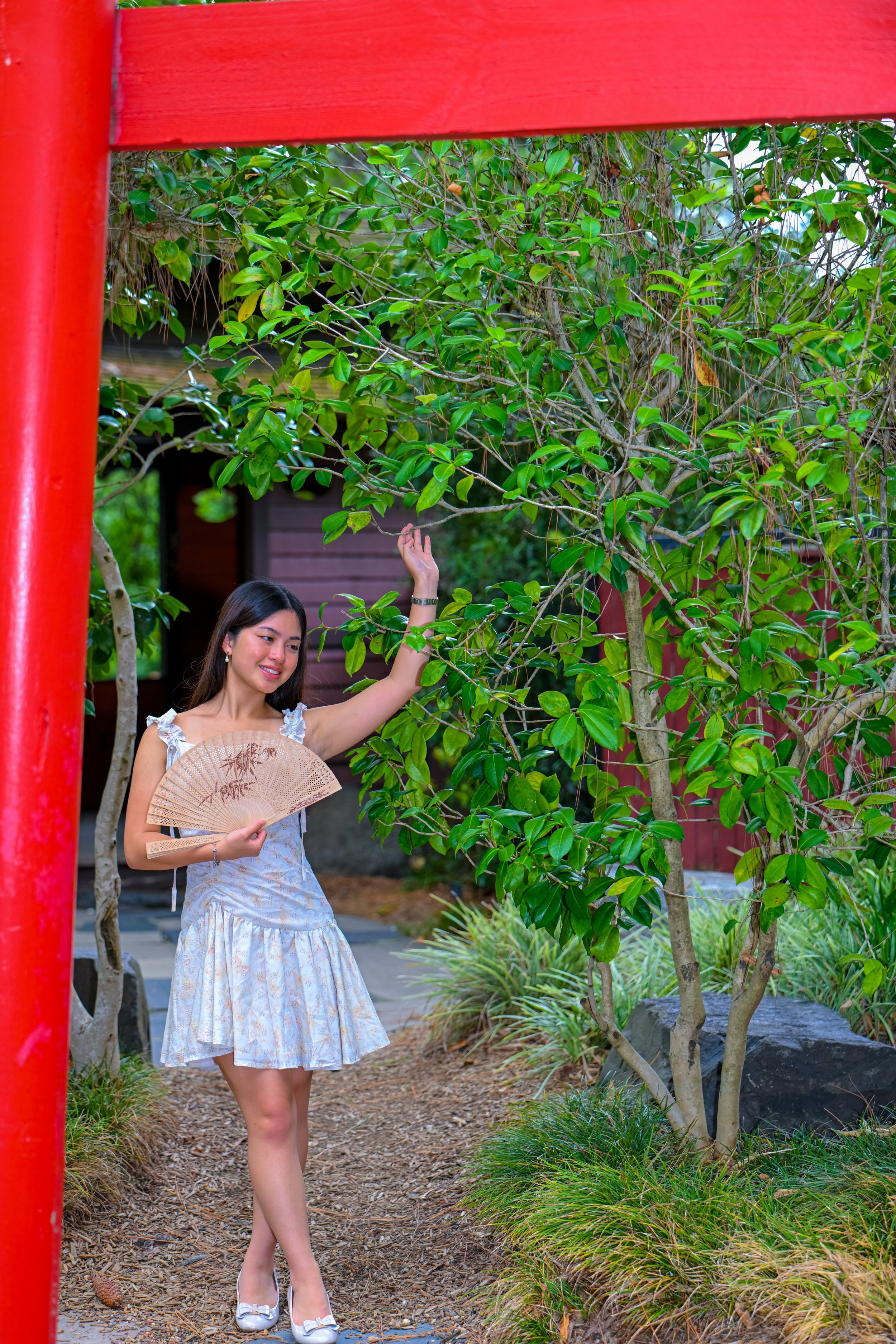 A woman in a light-colored dress holding a fan, standing on a garden path surrounded by trees and greenery, partially framed by a red structure.