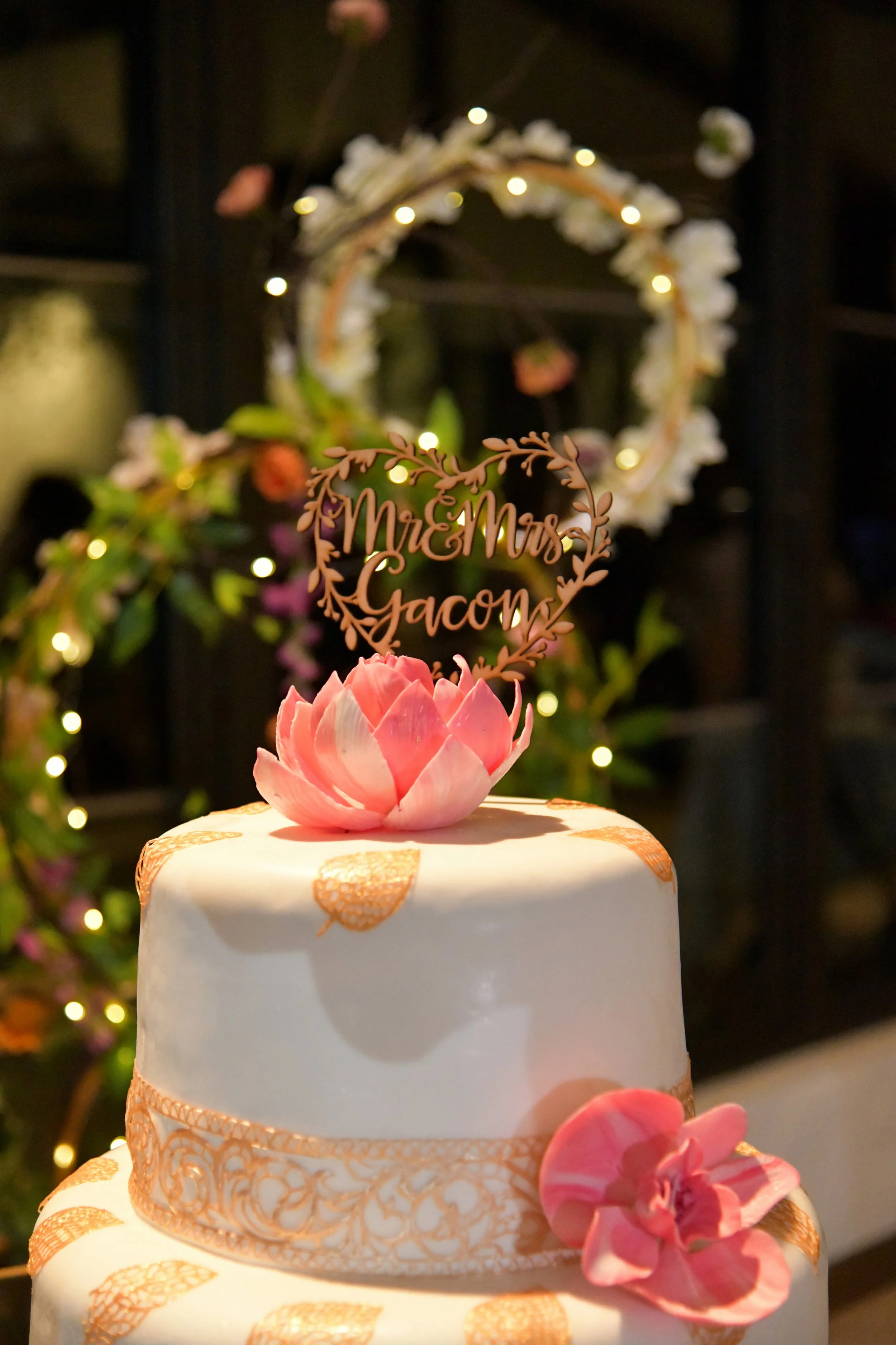 A wedding cake decorated with pink flowers and gold accents, with a topper that reads 'Mr & Mrs Gacon.' The background features floral and light decorations.