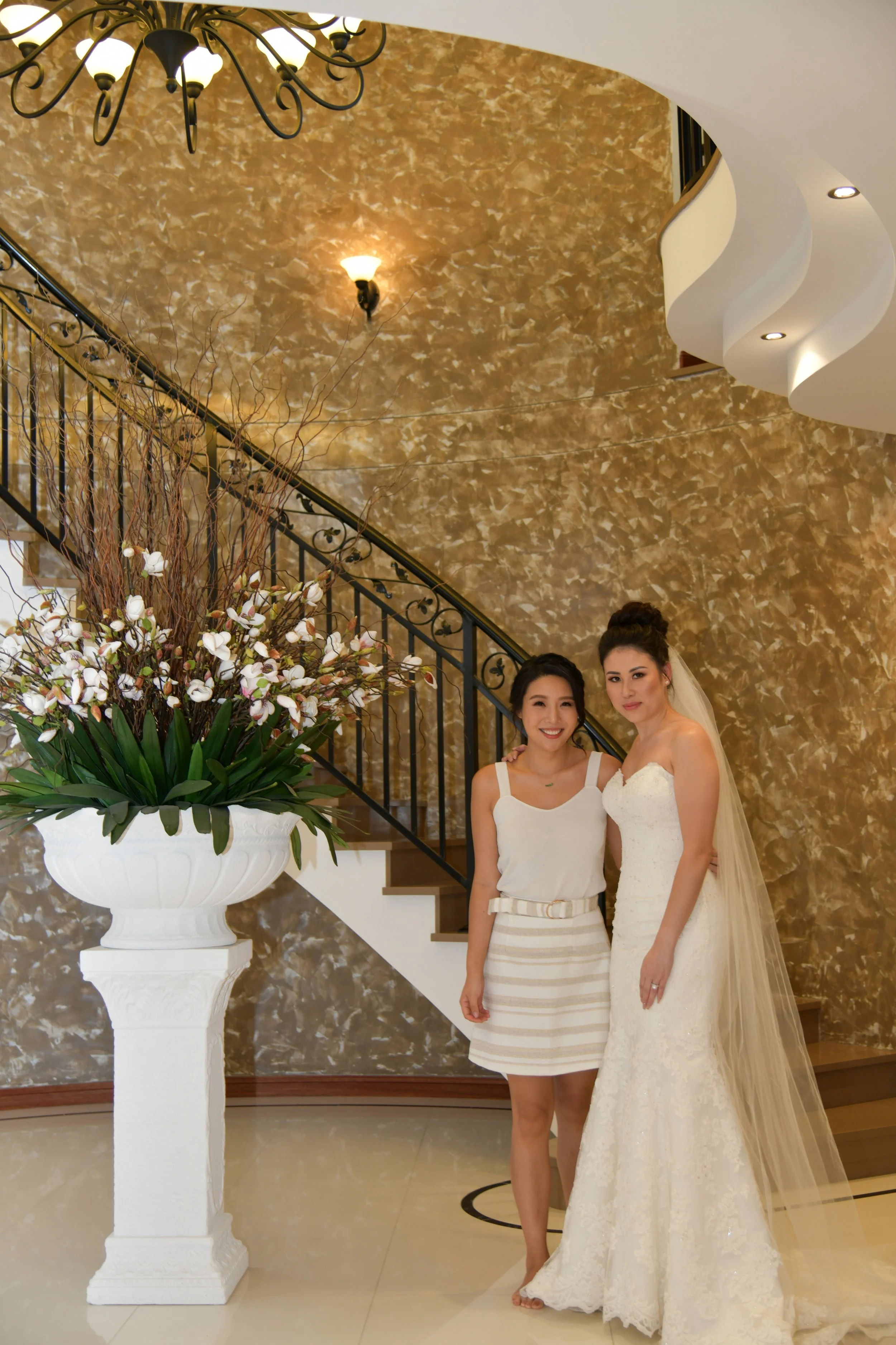 Two women standing by a staircase with a large flower arrangement in a white urn. One woman is dressed in a wedding gown with a veil, and the other is dressed in a casual white and beige striped skirt and top. They are smiling.