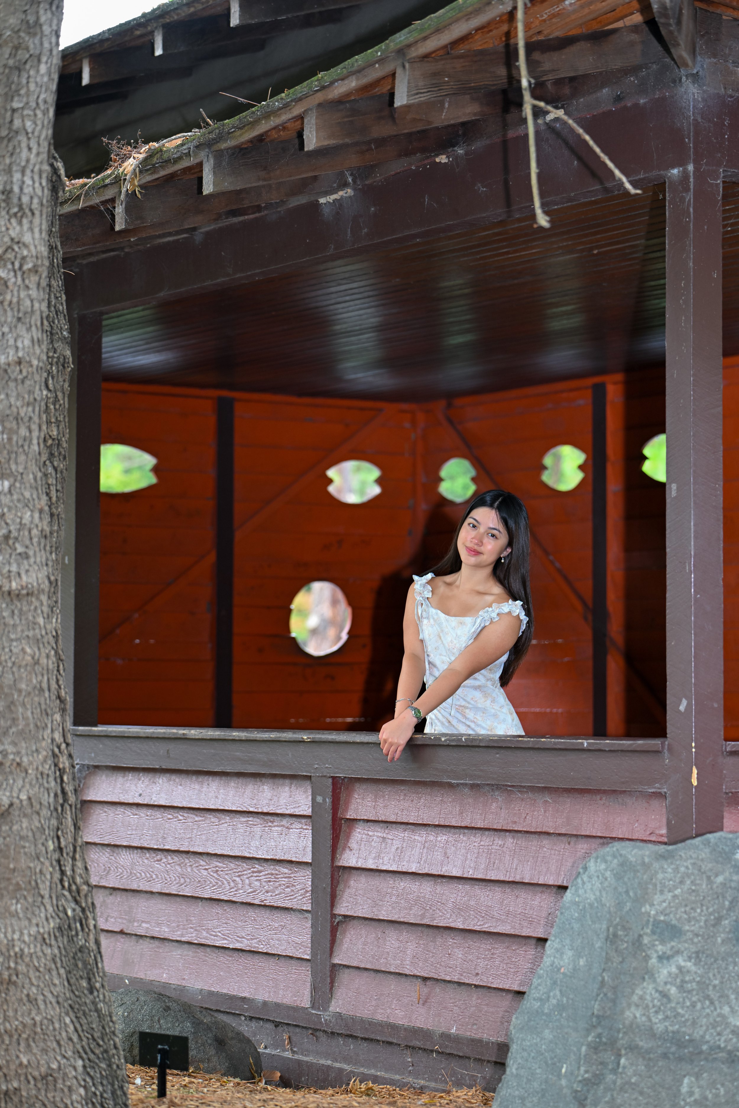 A young woman in a white dress standing on the porch of a wooden structure with circular cutouts, surrounded by trees and rocks.