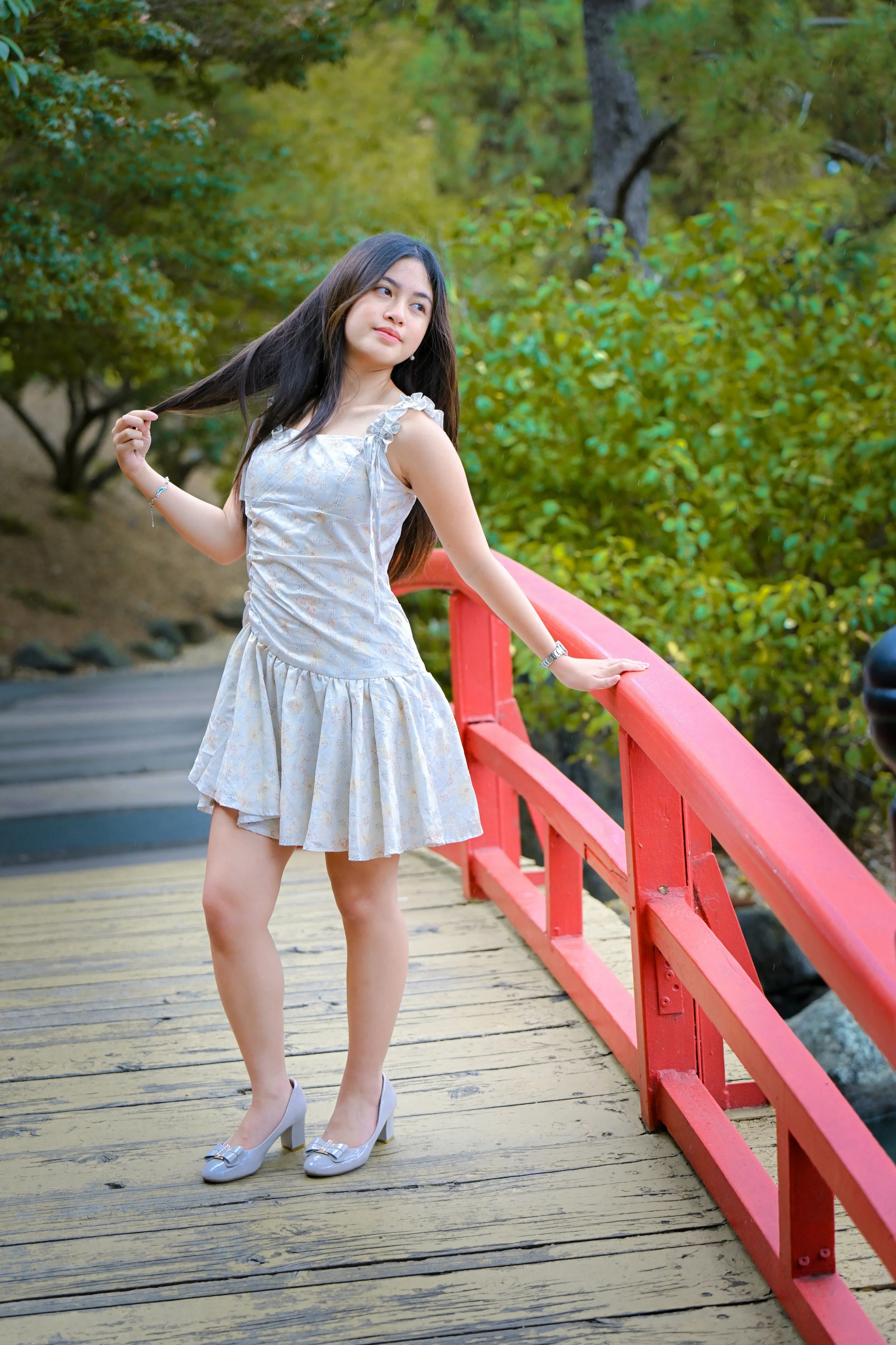 A young woman in a light-colored dress and gray high heels standing on a wooden bridge with red railings, holding her hair in one hand and resting the other hand on the railing. She is outdoors with green trees and foliage in the background.
