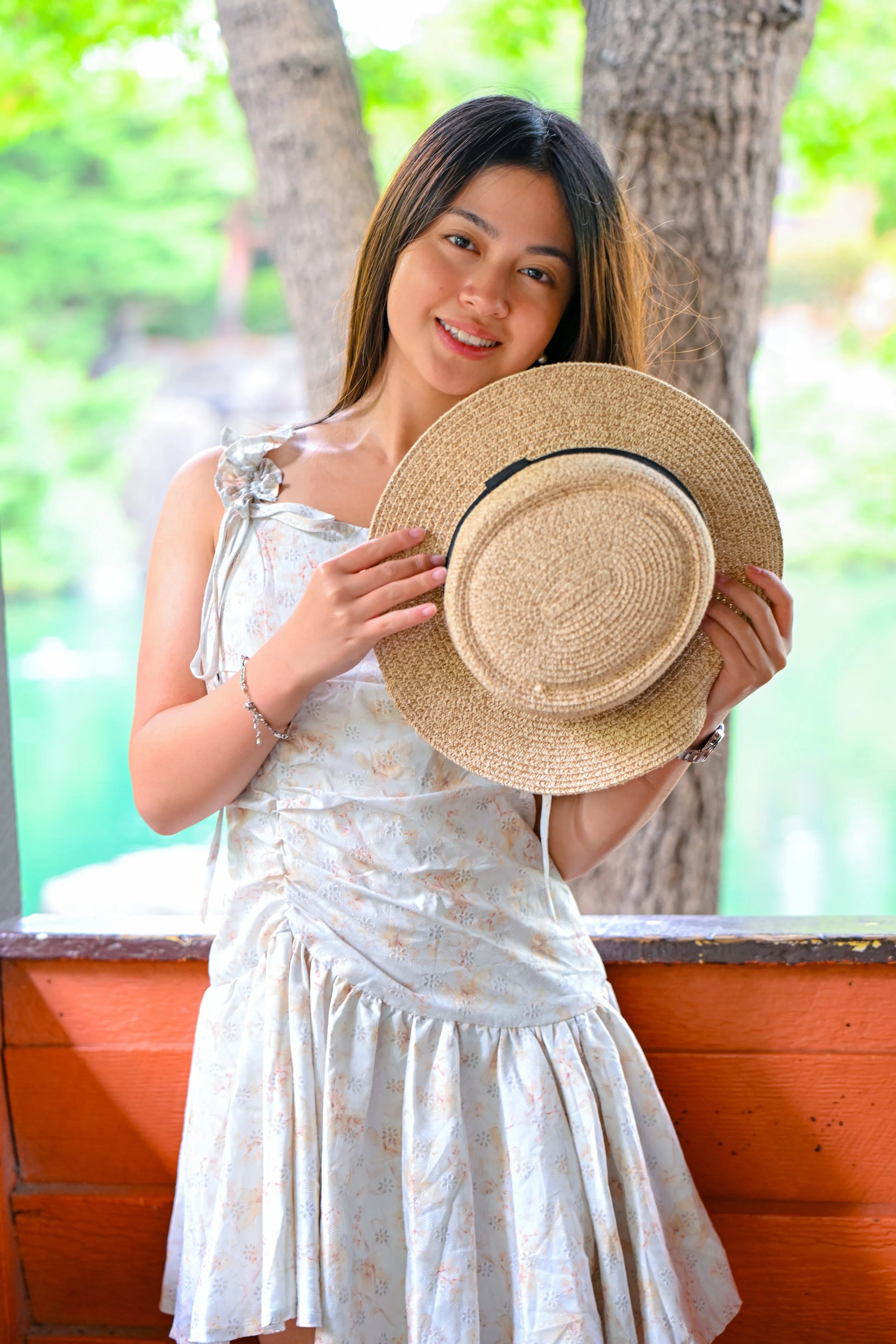A young woman with long brown hair holding a straw sunhat, standing outdoors by a lake with trees in the background.