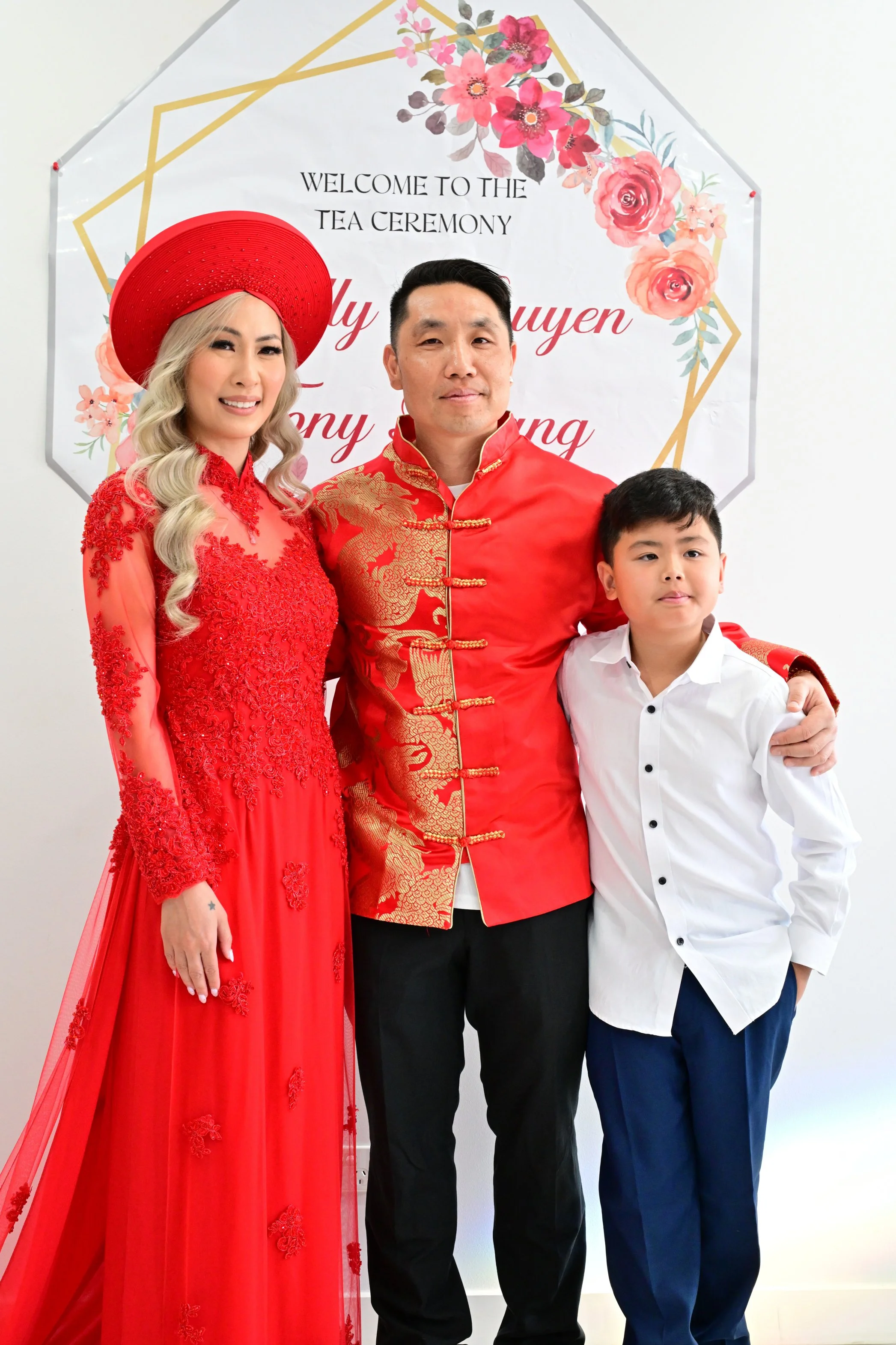 Family posing in traditional Vietnamese attire at a tea ceremony, with a floral welcome sign in the background.