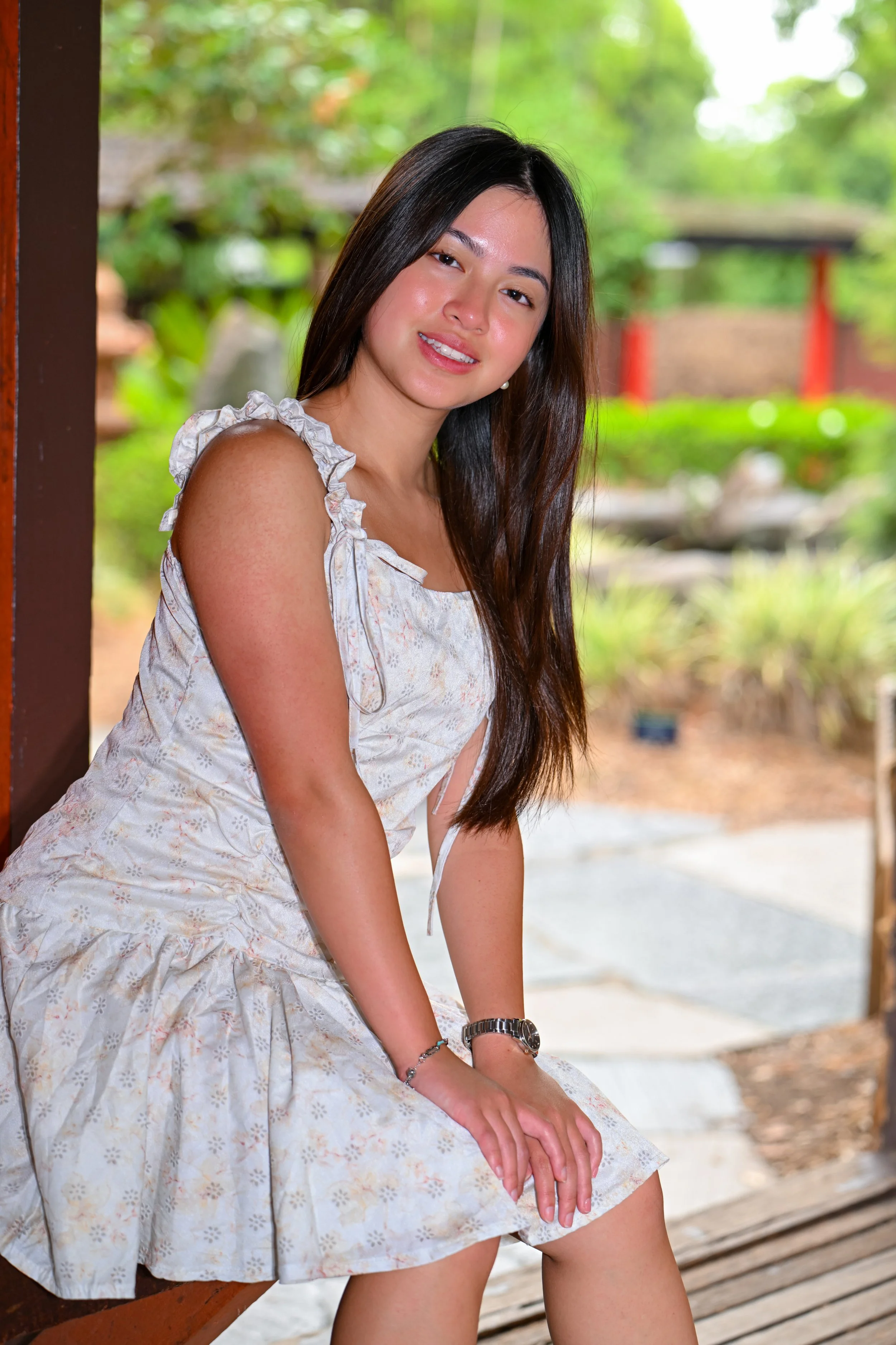 A young woman with long dark hair wearing a light-colored floral dress, sitting outdoors with a green garden background, smiling at the camera.