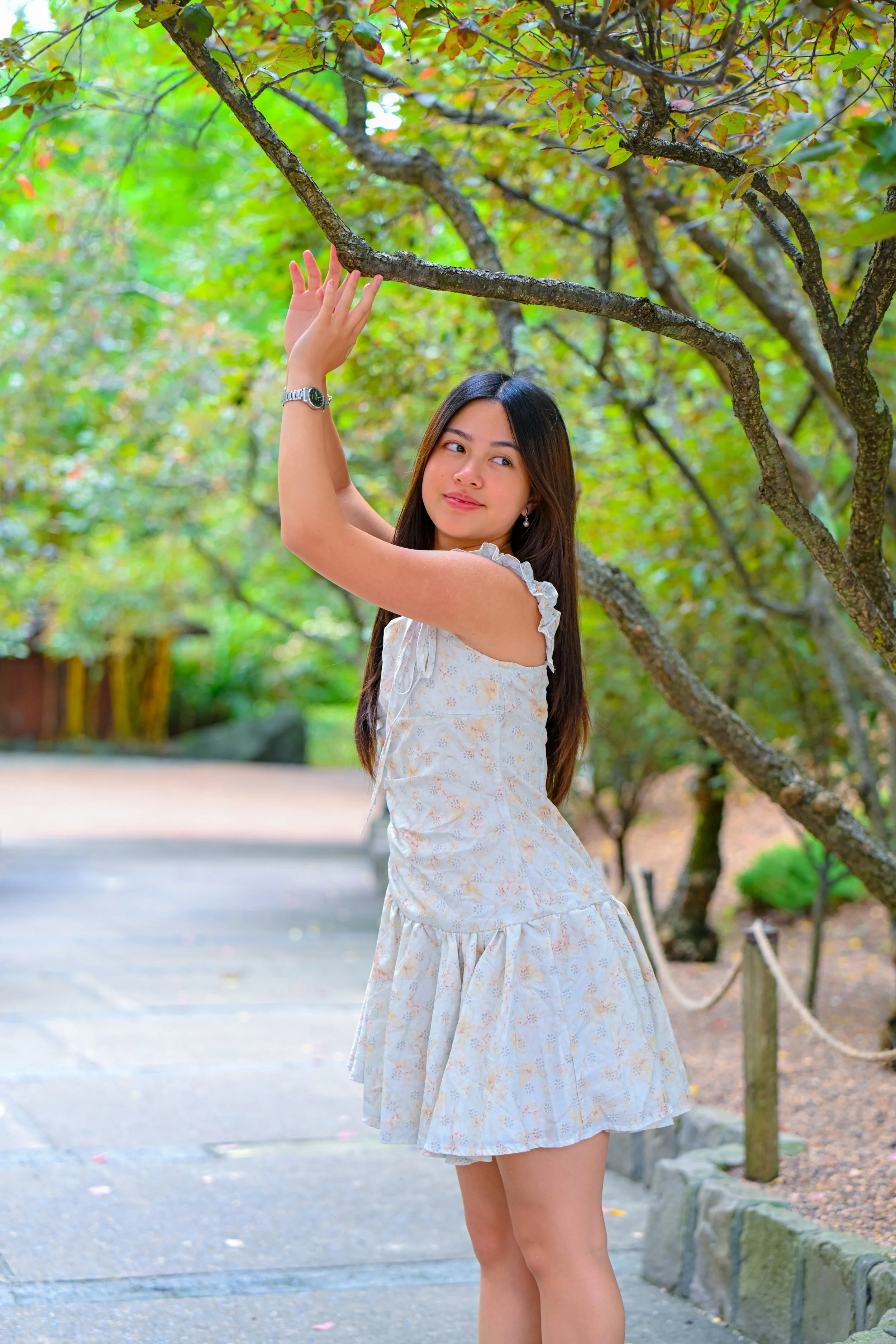 A young woman in a light floral dress standing on a park path, touching the branches of a tree with green and yellow leaves.