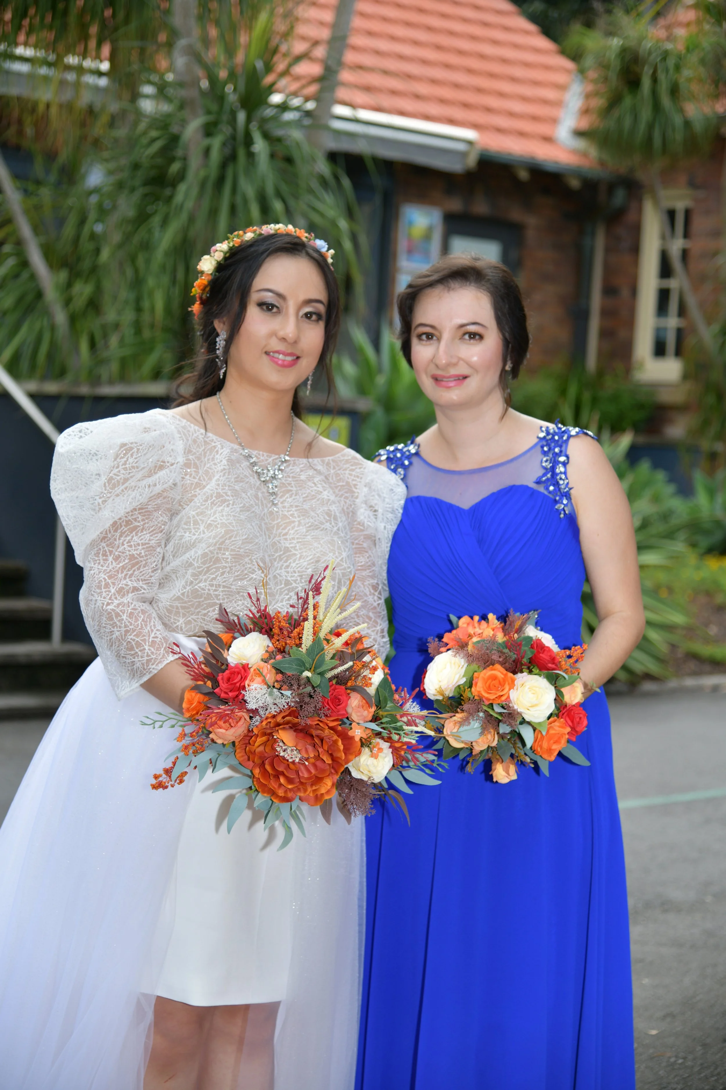 Two women standing outdoors, one in a white wedding dress with puffed sleeves and a floral crown, holding a large bouquet of orange, red, and white flowers. The other woman in a royal blue gown with embellishments on the shoulders, also holding a bou