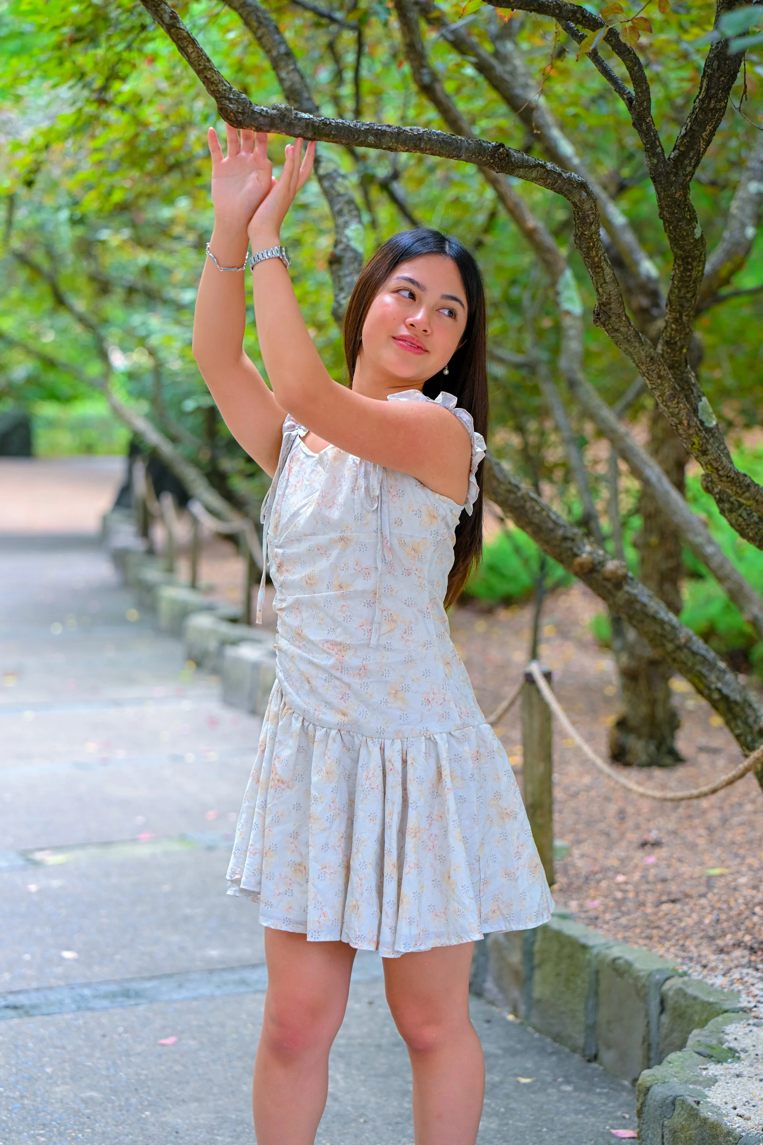 A young woman with long dark hair wearing a light-colored dress with floral patterns, standing on a pathway in a park, holding an overhead branch of a tree with both hands, surrounded by green trees and foliage.