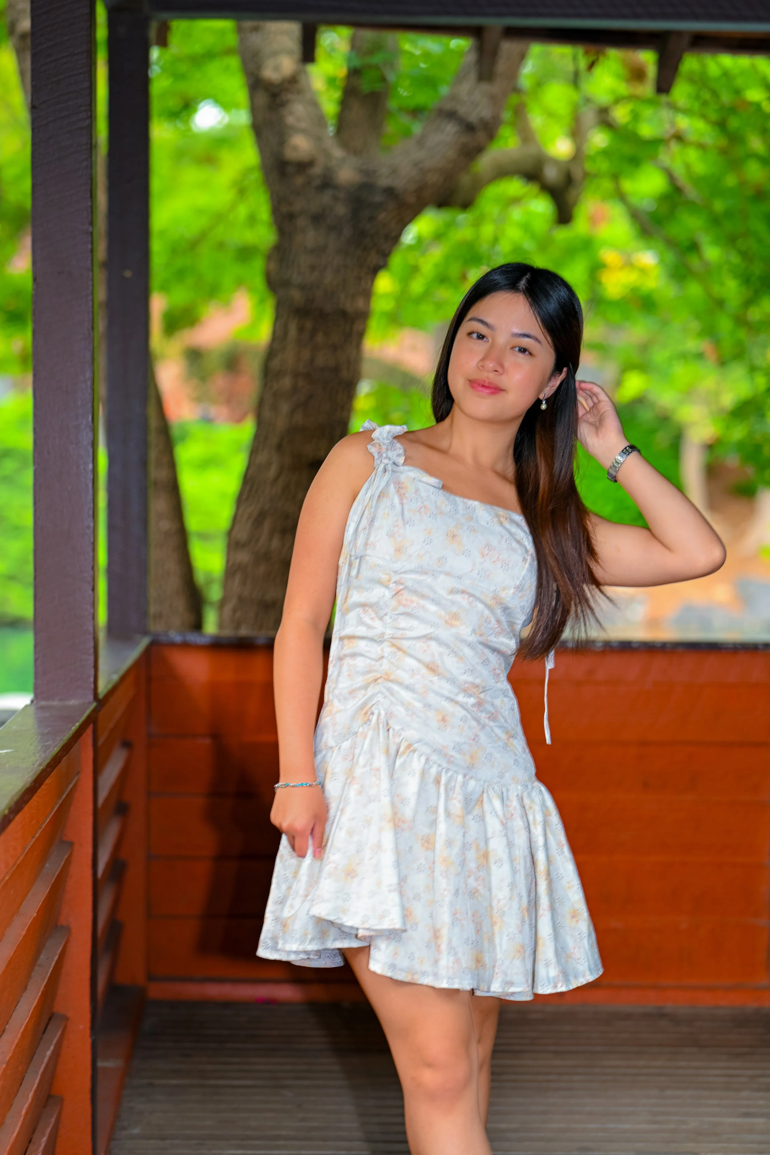 A young woman with long dark hair standing on a wooden patio, wearing a sleeveless light-colored floral dress, with green trees in the background.