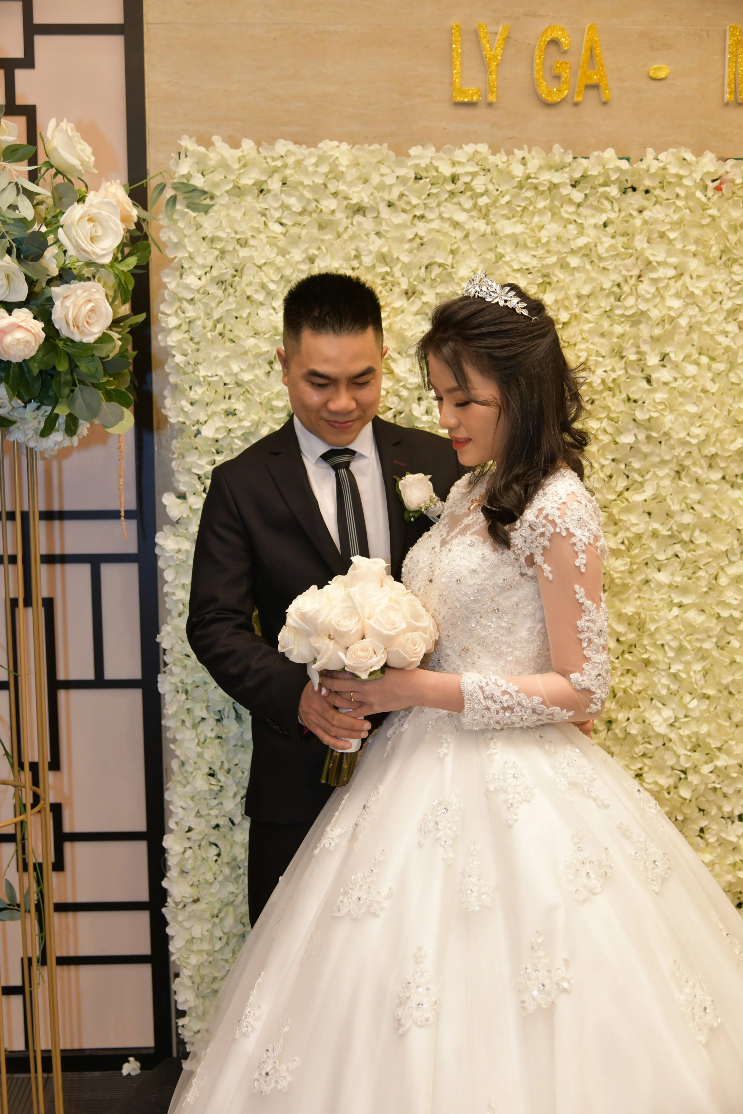 A bride in a white wedding gown and a groom in a black suit with a tie, holding a bouquet of white roses, standing in front of a floral backdrop with the text "LY GA" in gold letters.