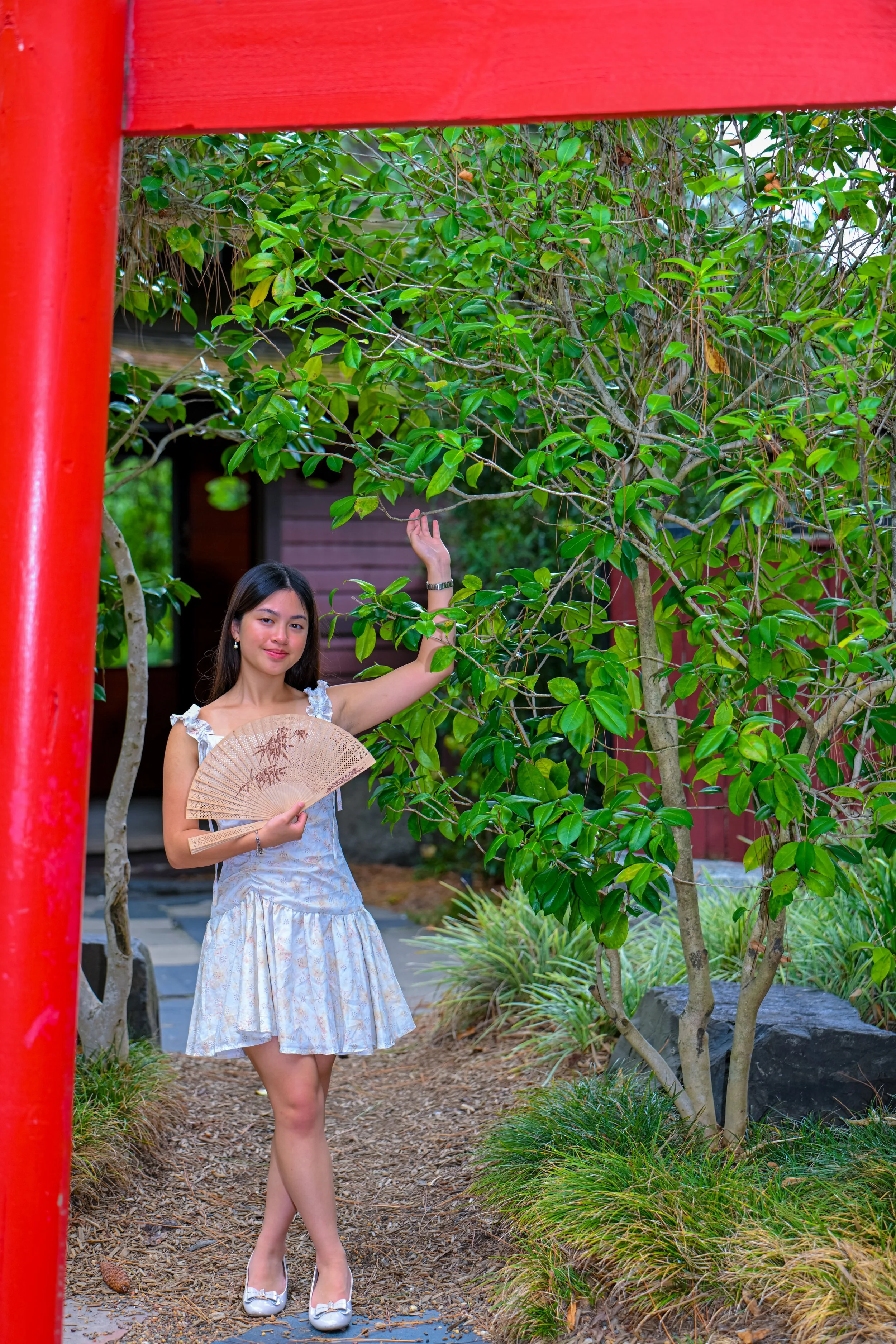 A woman in a light-colored dress holding a fan and touching a tree branch in a garden with greenery and a red structure.