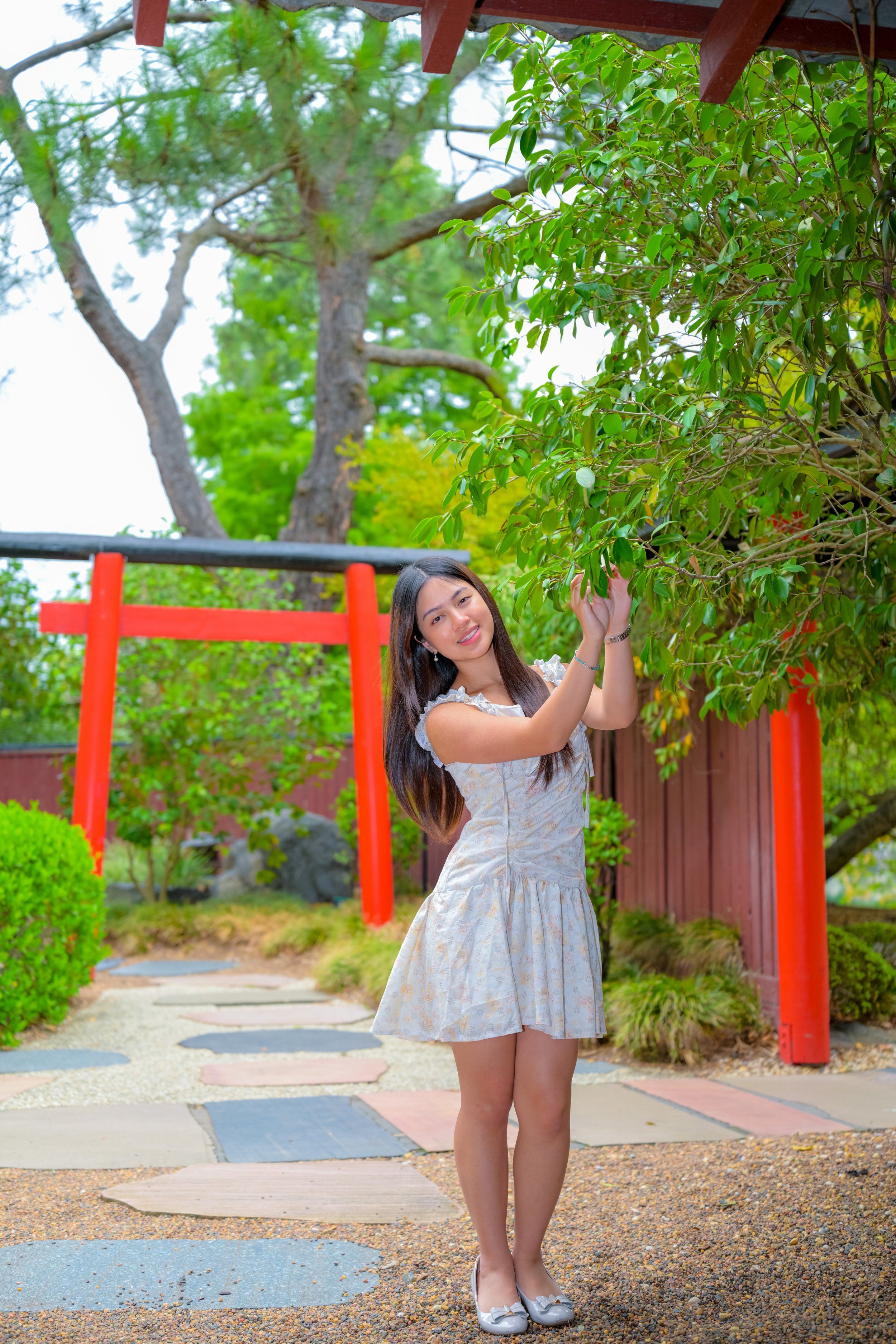 A young woman in a light-colored dress standing on a stone path in a garden with a red torii gate and green trees.