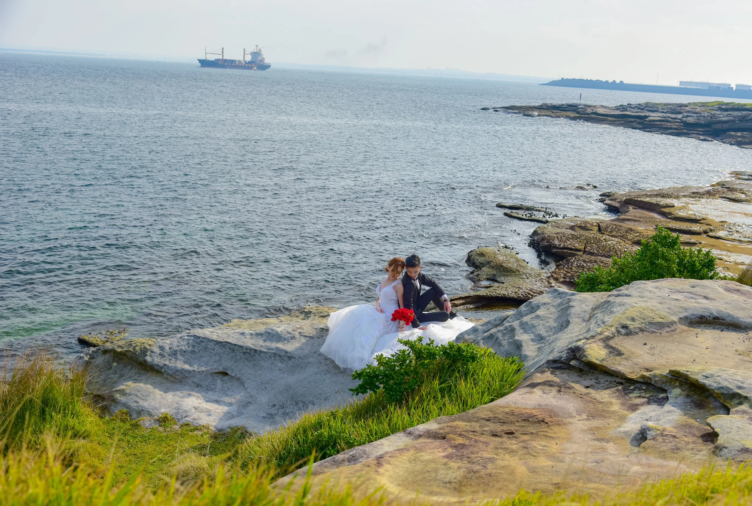 A couple dressed in wedding attire sitting on rocks by the sea, with a large cargo ship in the distance.