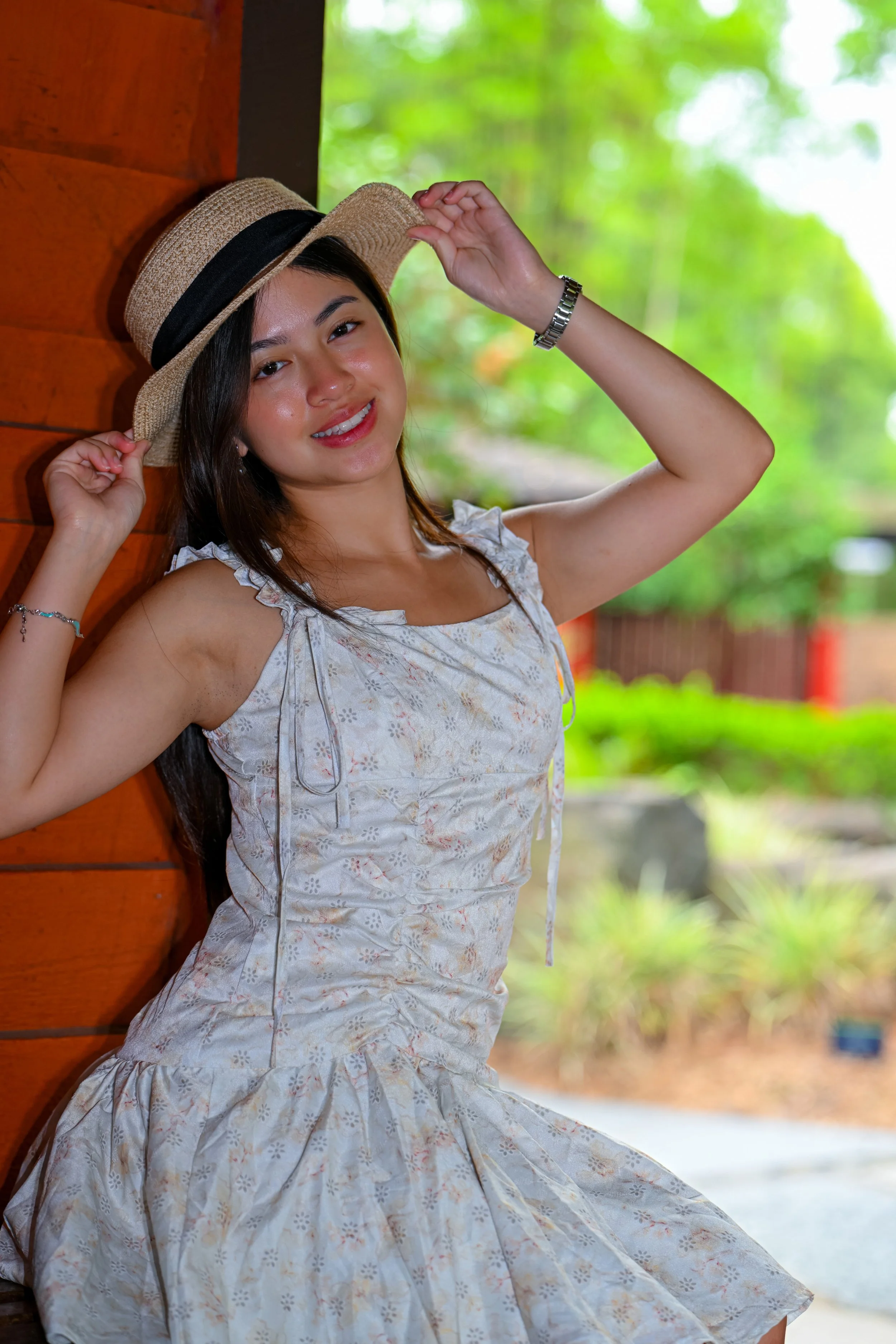 A young woman with long dark hair smiling, wearing a white dress with lace details and holding a straw hat with a black ribbon, leaning against a wooden wall outdoors with green foliage in the background.