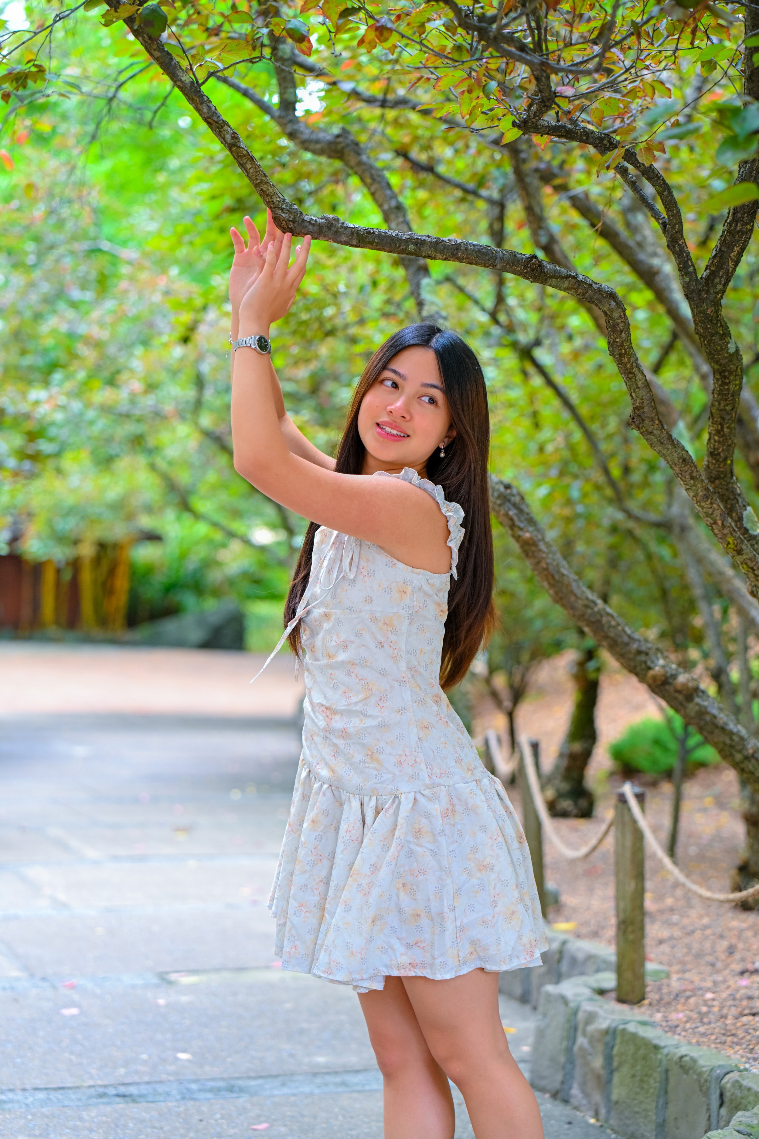 A young woman with long dark hair standing outdoors in a park, touching a tree branch with a gentle smile on her face. She is wearing a light-colored, sleeveless dress with ruffled shoulders.