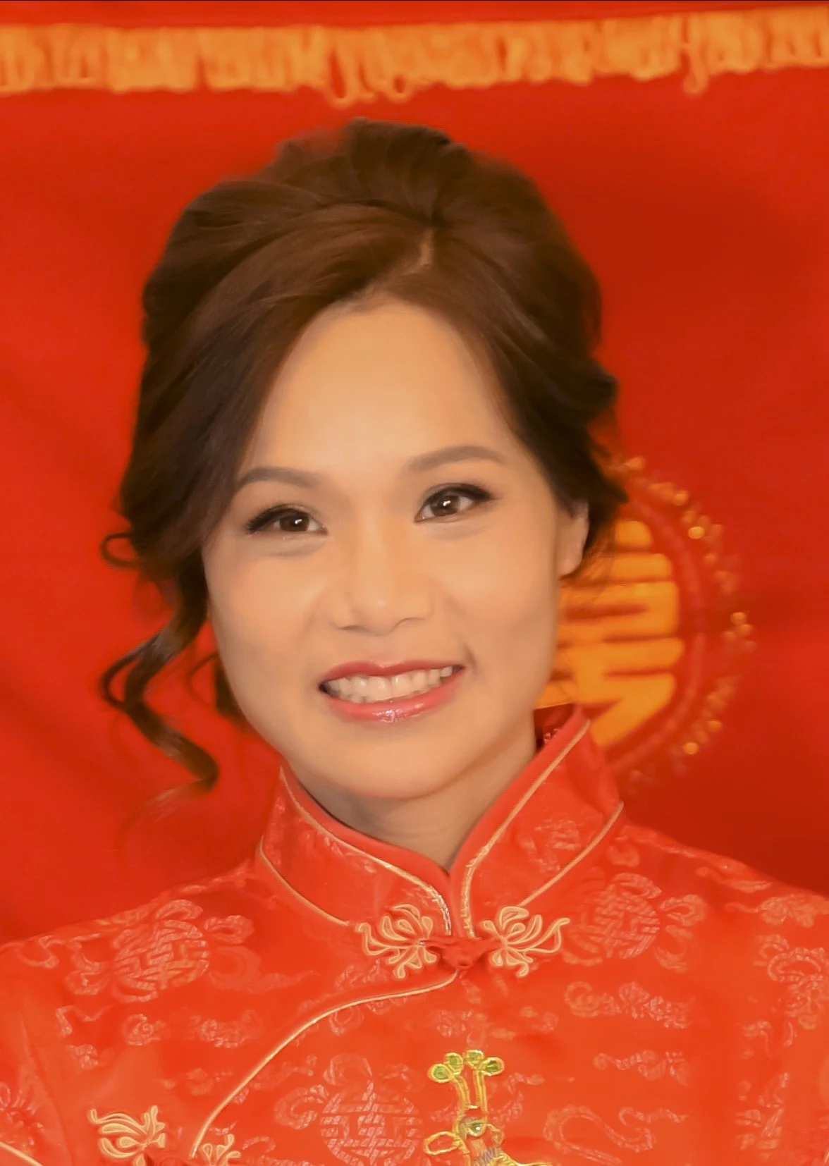 A woman wearing a traditional red Chinese dress with gold embroidery, smiling with her hair styled in loose curls, against a red background.