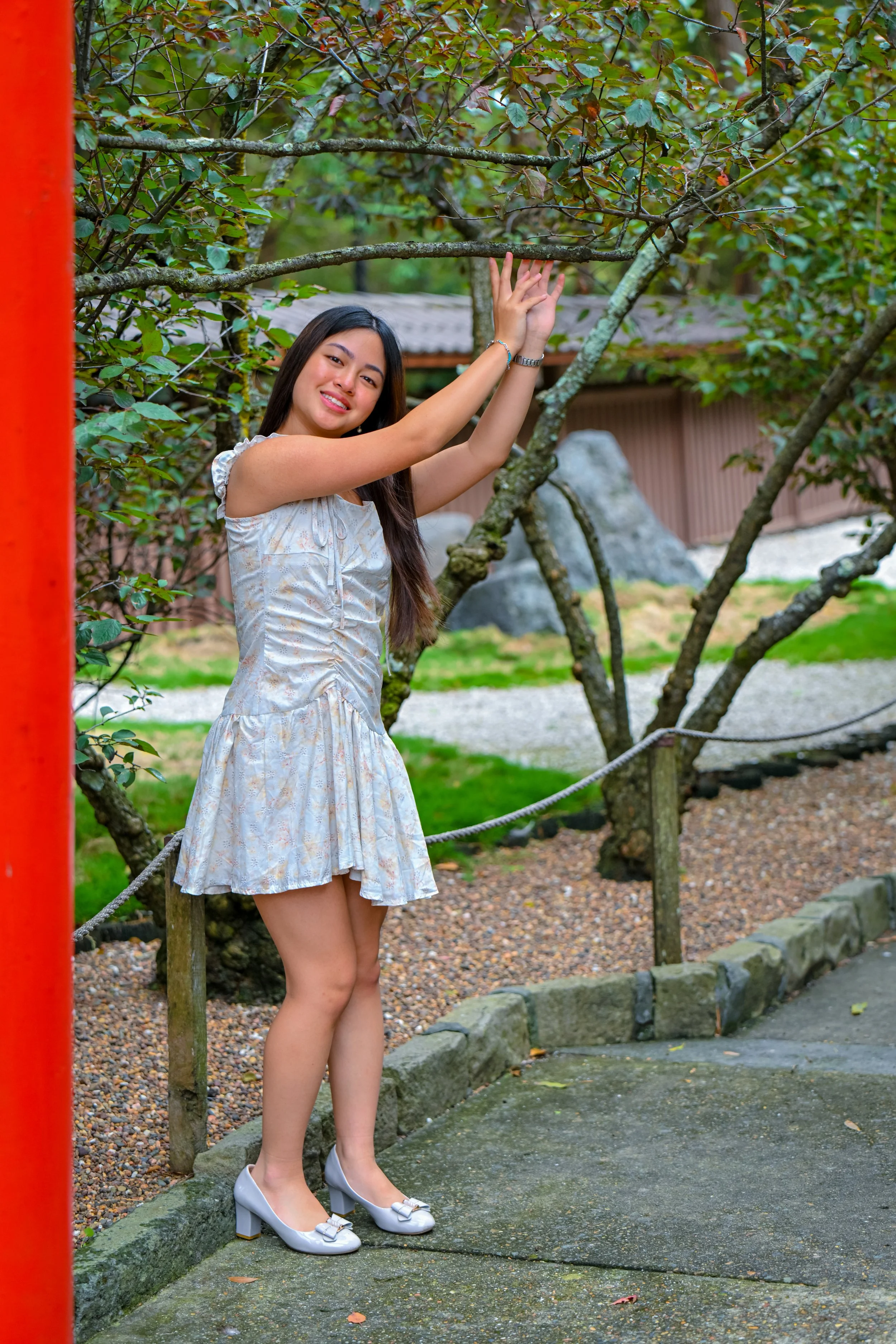 A young woman with long dark hair, wearing a light-colored dress and silver shoes, stands outdoors beside a tree, smiling and holding onto a branch.