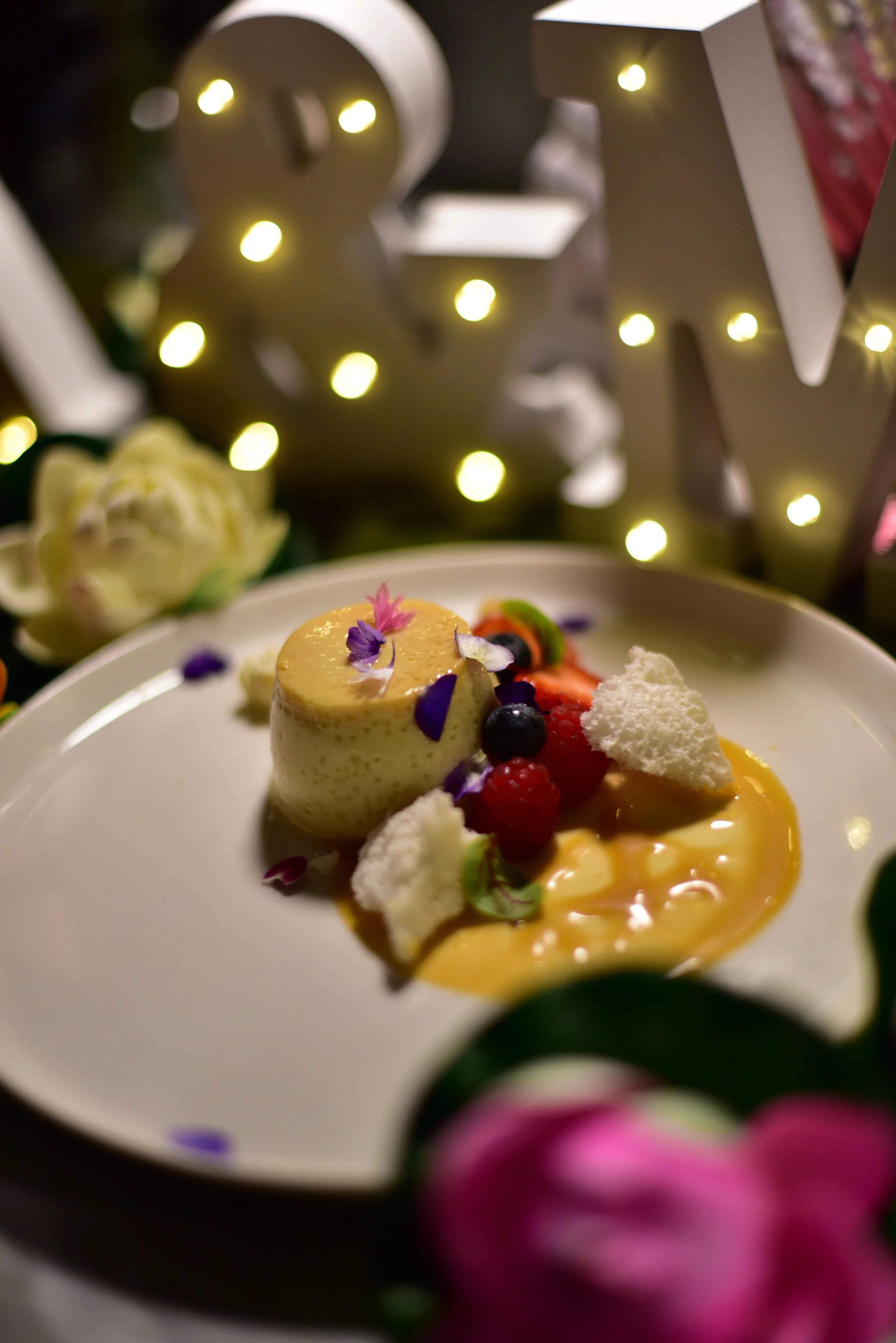 A dessert plate with a small round mousse garnished with flower petals, surrounded by berries, meringue pieces, and sauce, with decorative lights and flowers in the background.