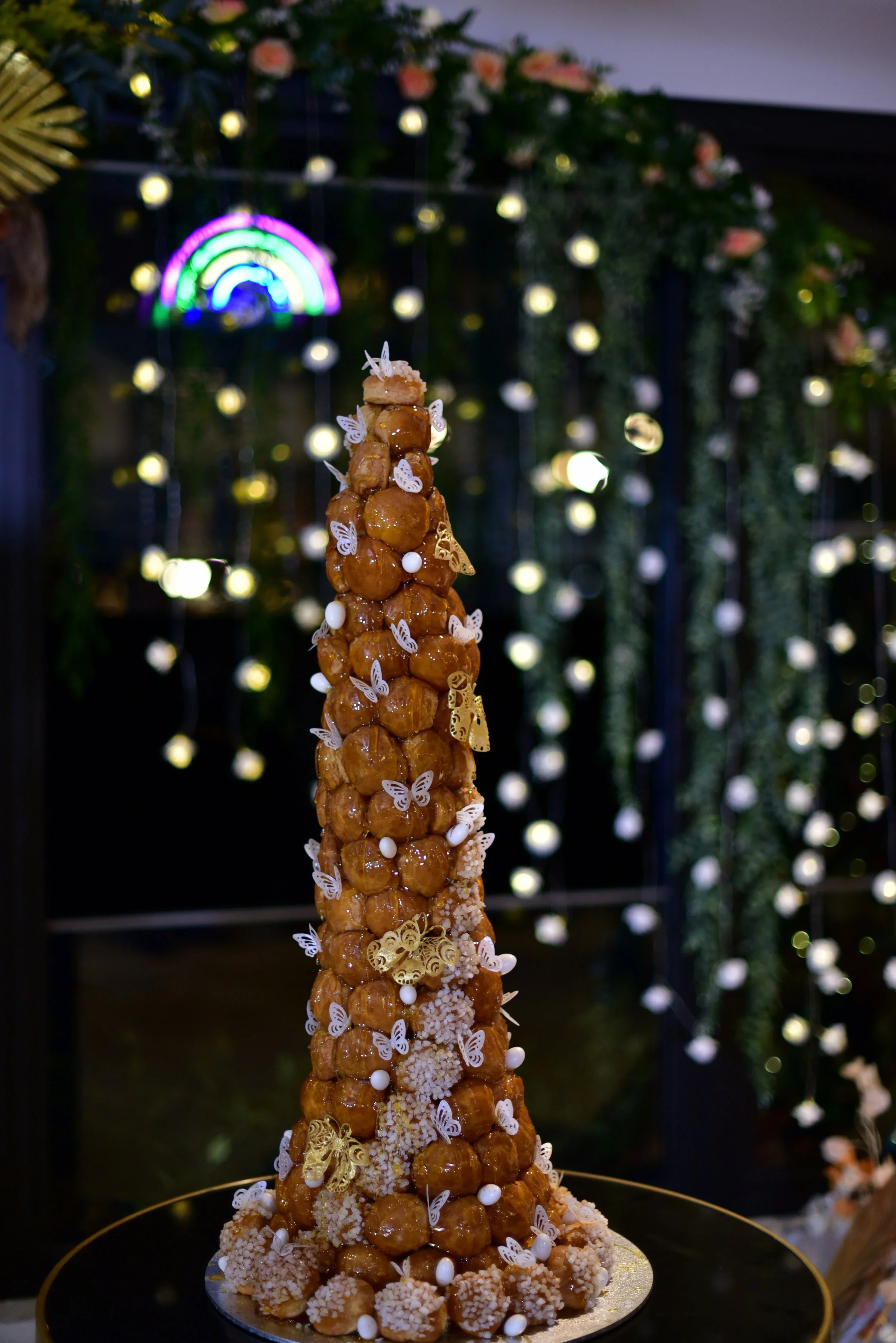 A towering croquembouche decorated with white butterflies, gold and white candies, and floral accents, set against a background of bright lights and greenery.