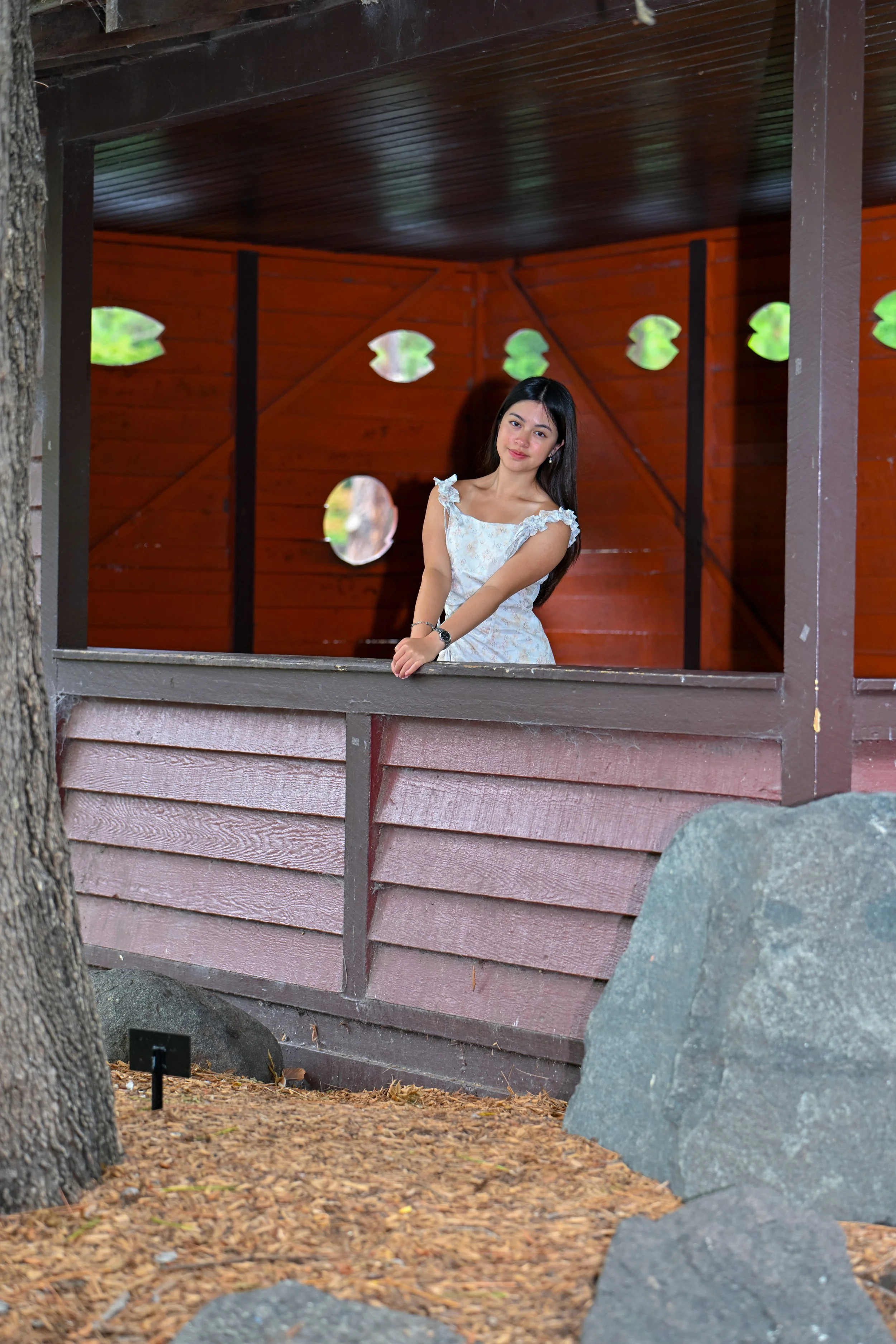 Young woman in white dress leaning on the railing of a wooden porch with circular cutouts, outdoors.