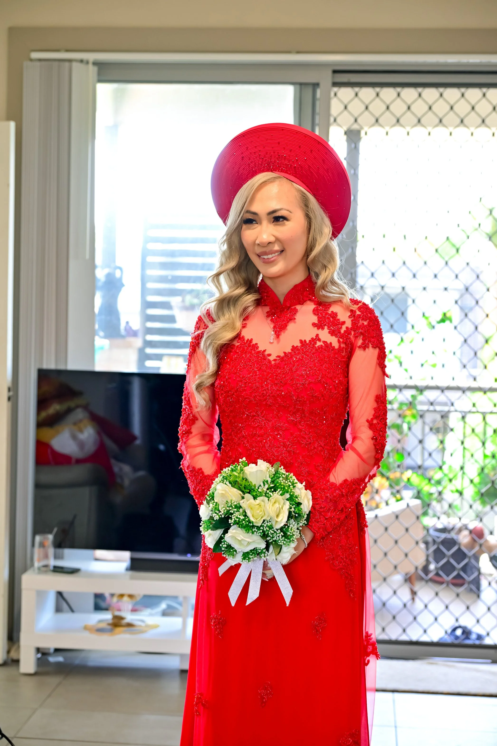 A woman in a red lace wedding dress holding a bouquet of white roses, wearing a large red hat, standing indoors near a sliding glass door.