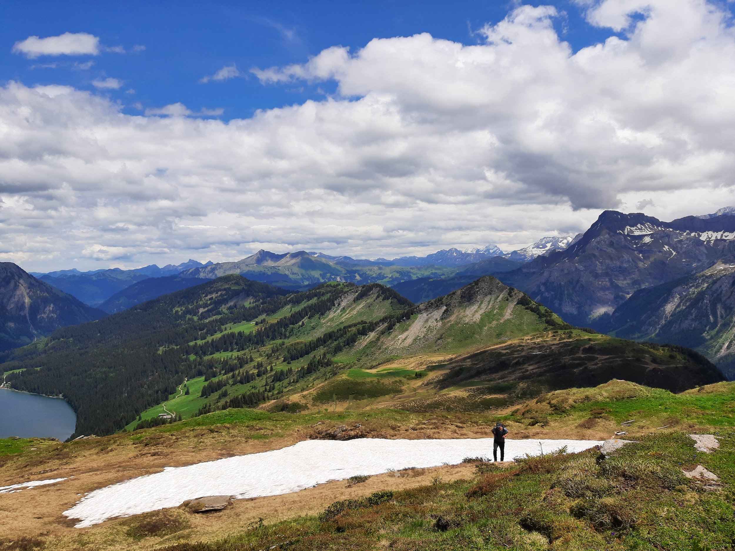 Mountain landscape with green hills, snow-capped peaks in the distance, a lake, and a person standing near a patch of snow.
