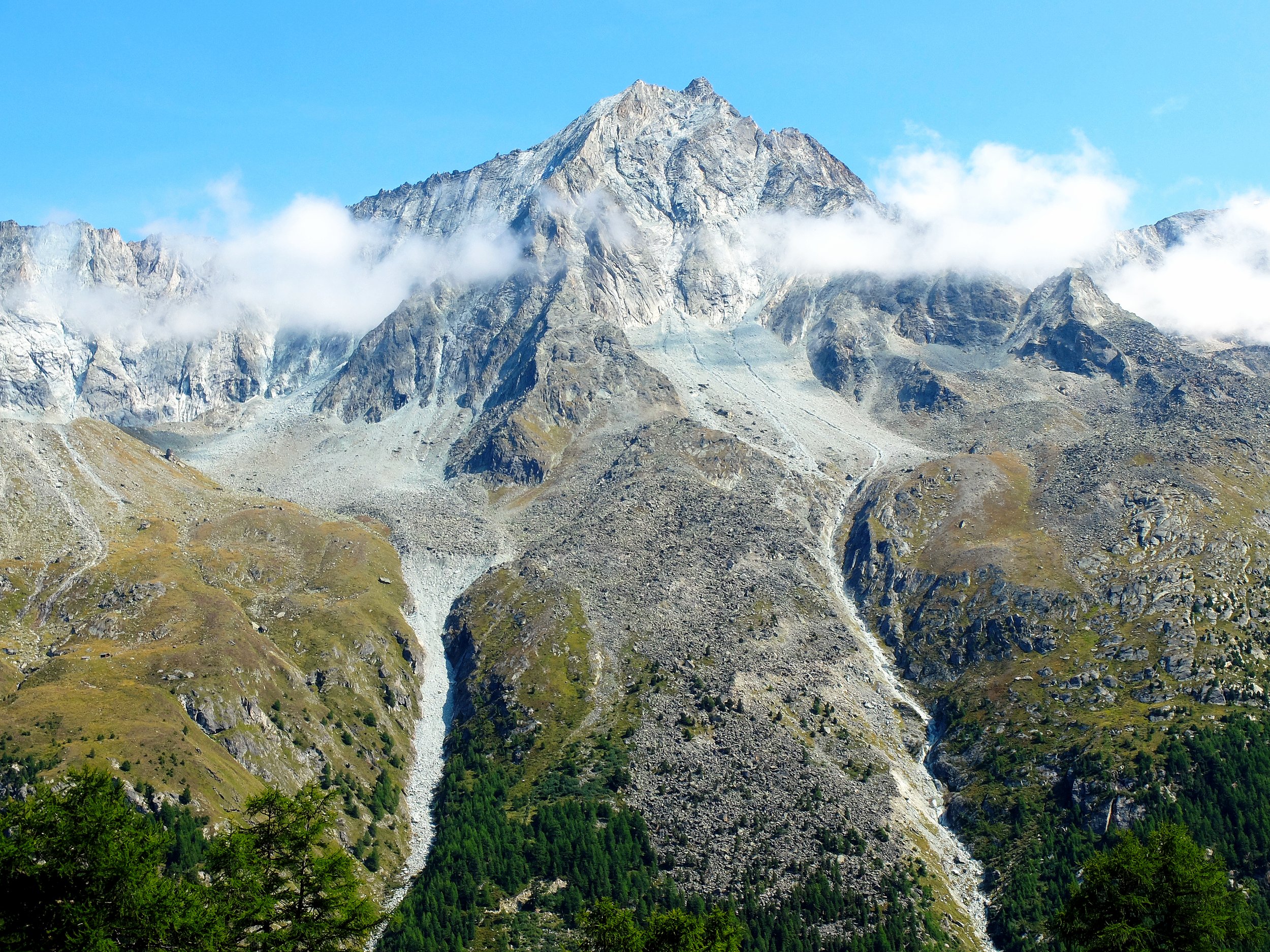 Mountain landscape with rocky peaks, green forest at the base, and clouds surrounding the summit.