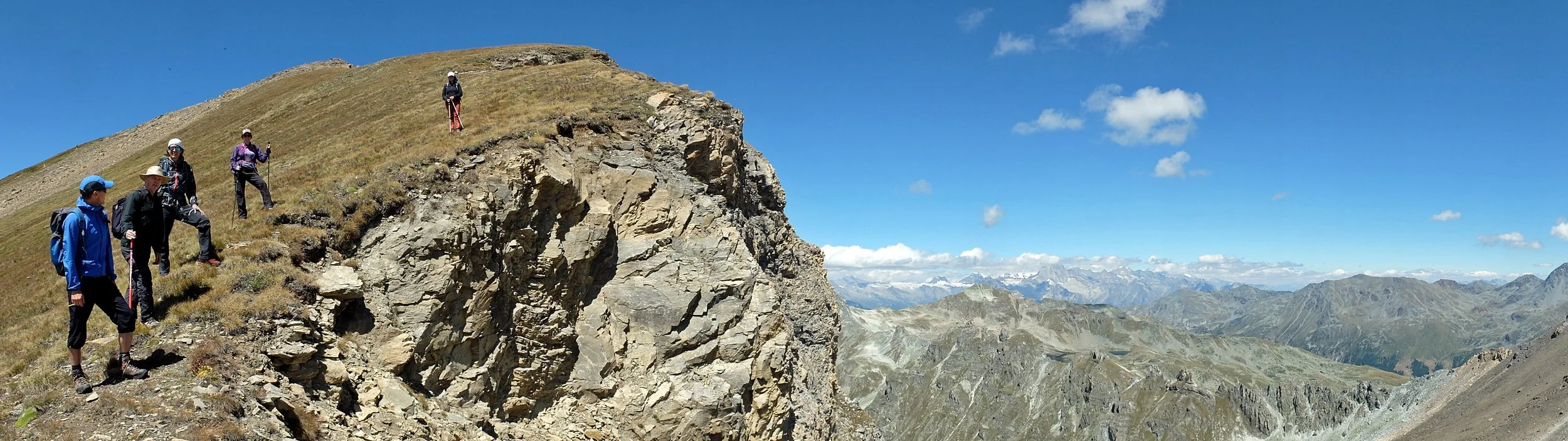 Group of hikers walking along a mountain trail with rocky cliffs, surrounded by mountain ranges and a blue sky.