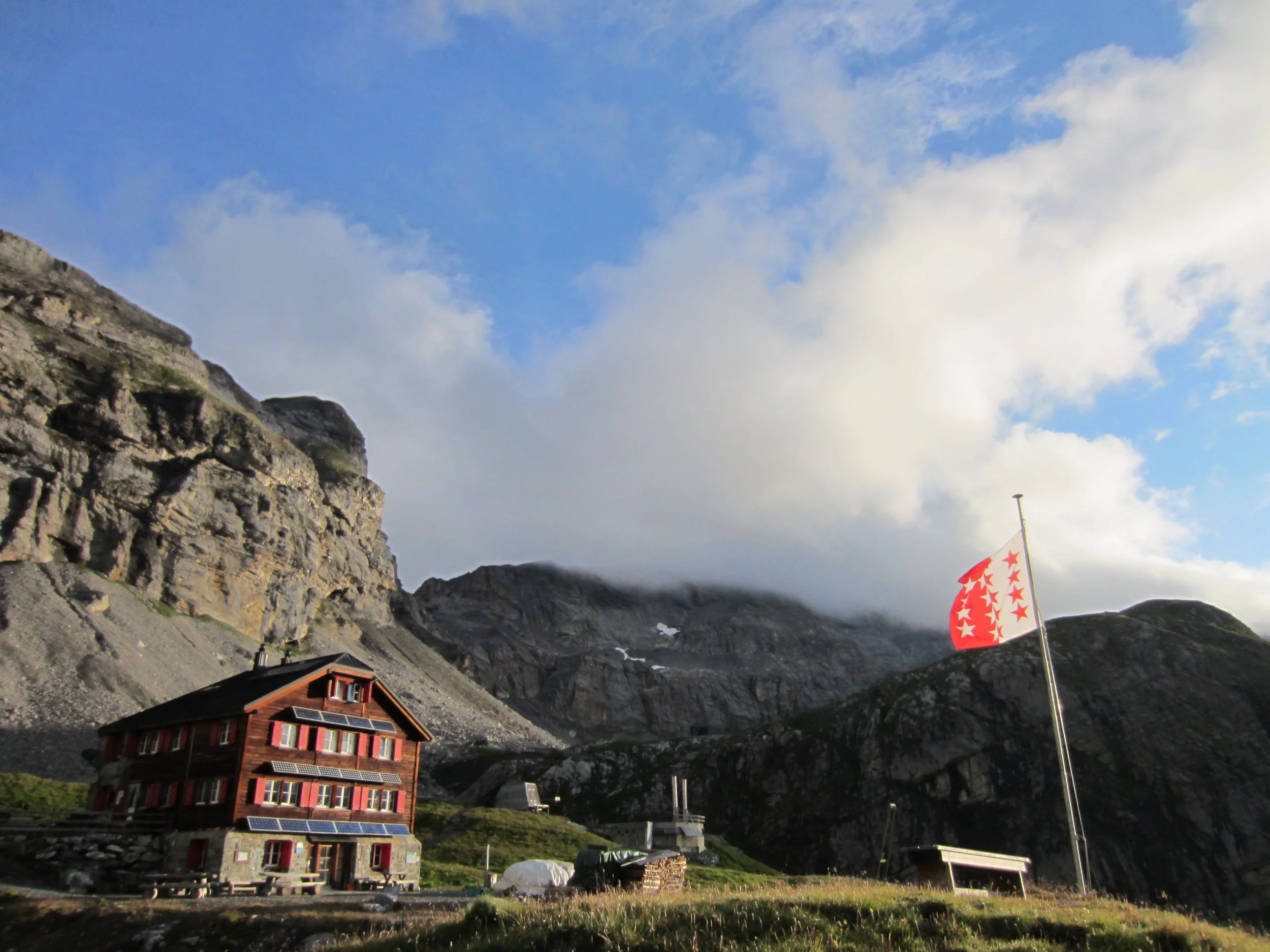 A rustic multi-story cabin with red and black shutters and solar panels, situated in a mountainous area with rugged rocks and a partly cloudy sky. A flag with red stars on a white background is on a pole nearby.