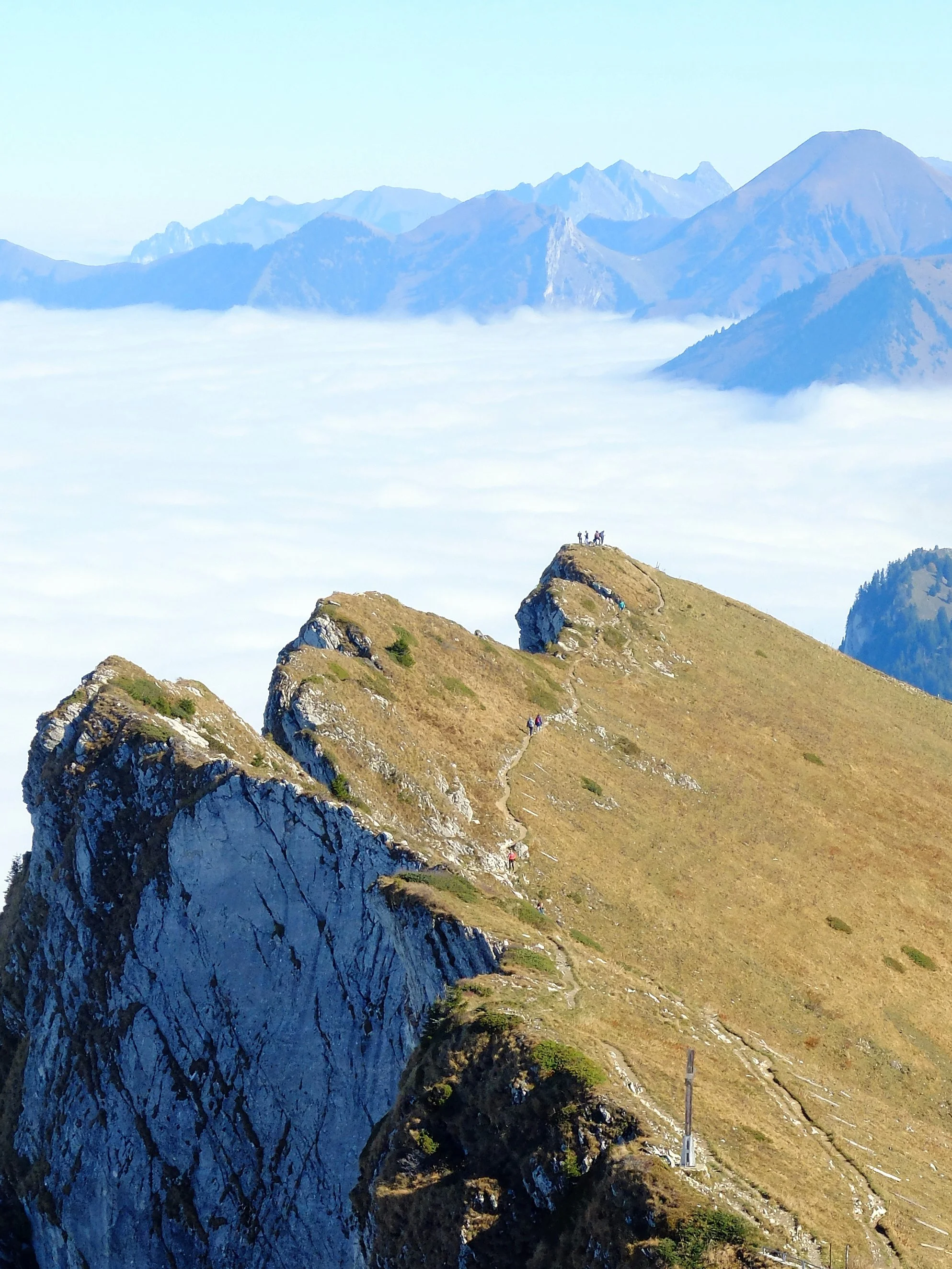 Mountains with a trail leading to a peak; several hikers on the trail and at the summit; clouds below the mountain ridges; distant mountain peaks in the background.