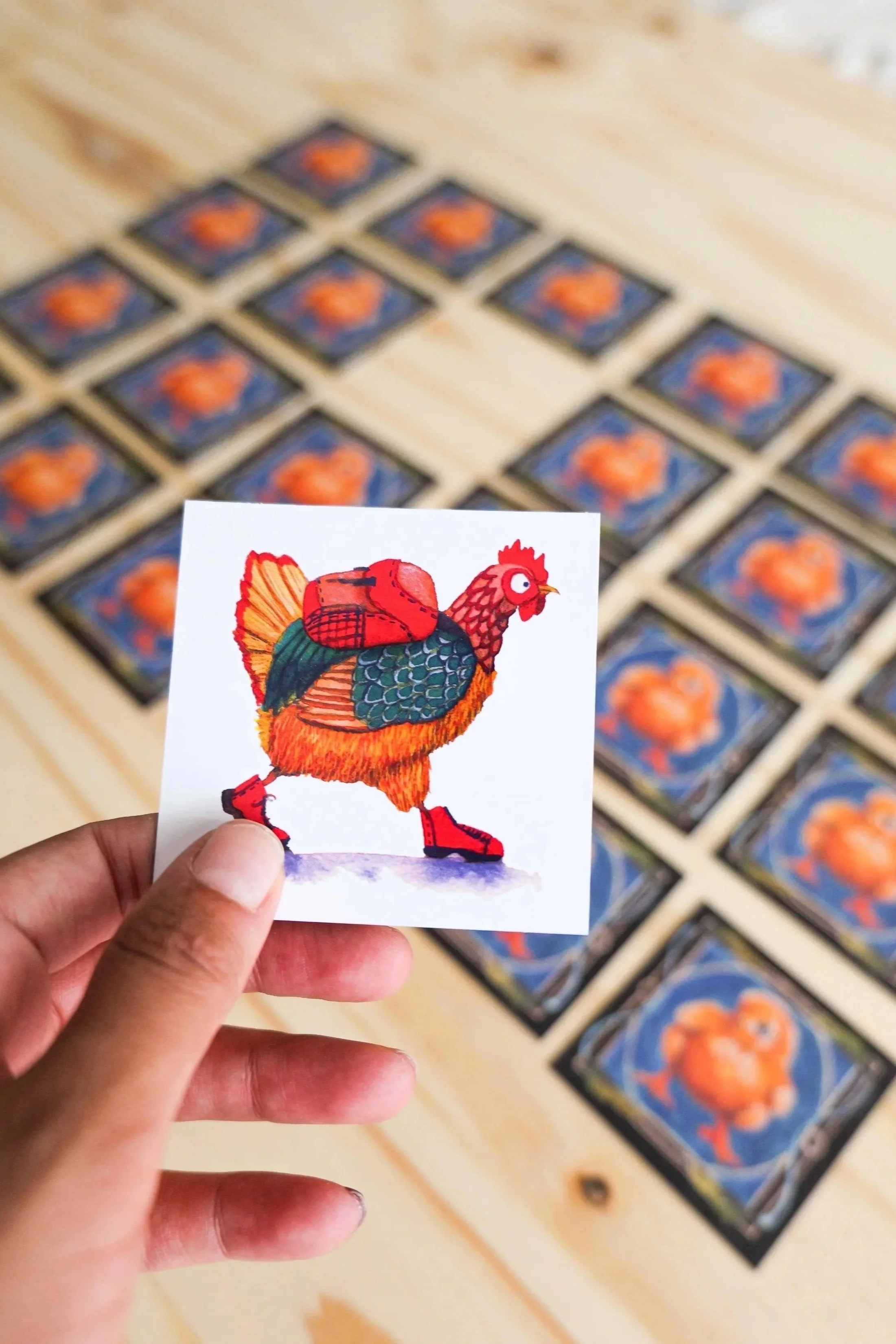 Une main tient une carte du jeux de memory avec une illustration d'une poule colorée portant un sac à dos et des chaussures de sport, devant plusieurs autres cartes similaires étalées sur une surface en bois. 