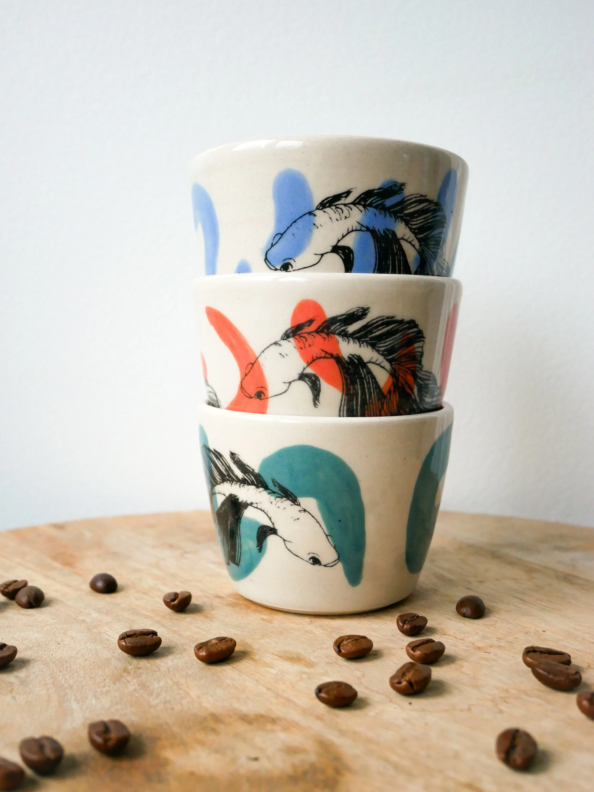 Trois tasses de céramique artisanale empilées avec des dessins de poissons, placées sur une table en bois, entourées de grains de café.