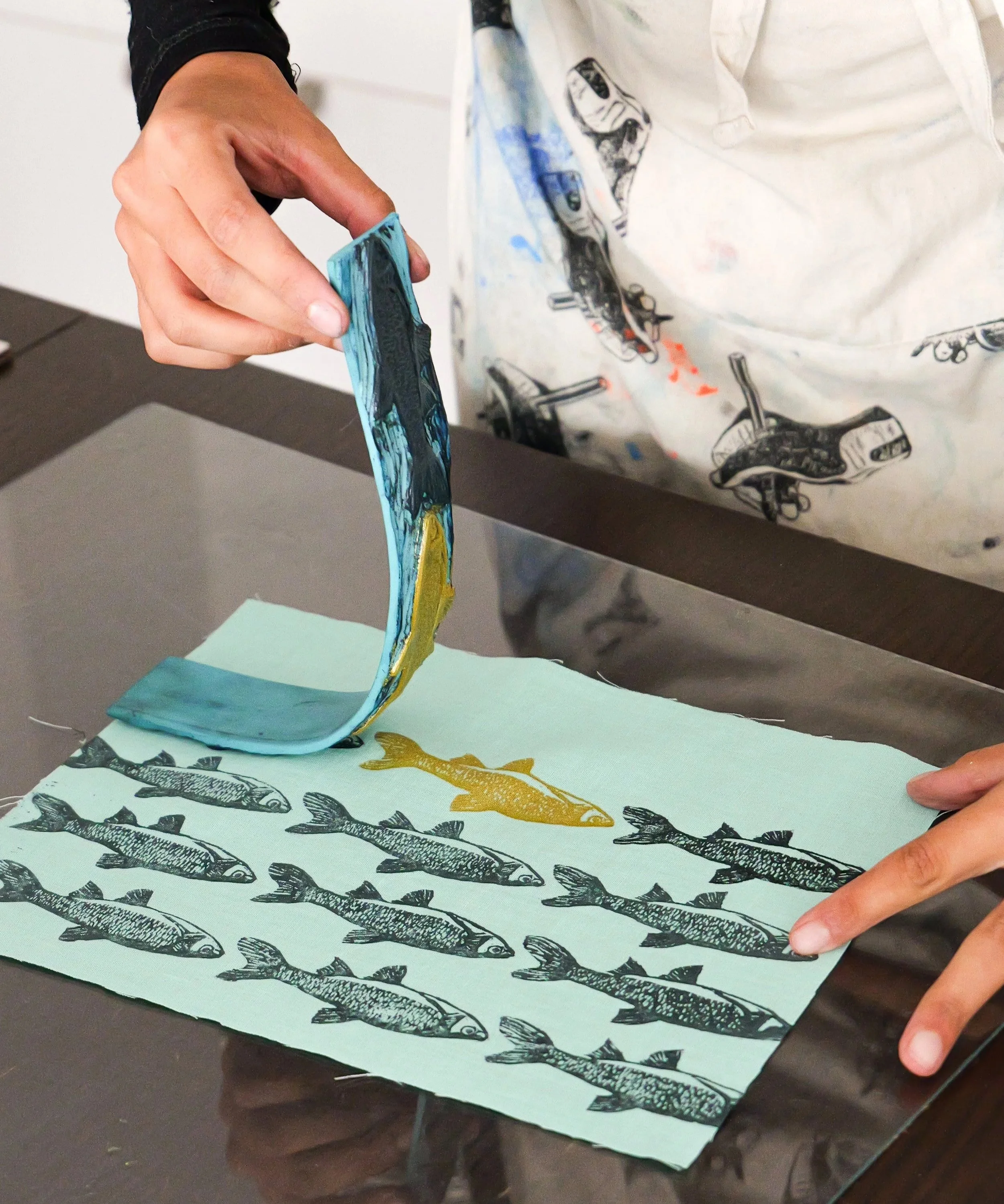 Une personne qui fait de la linogravure sur du textile de seconde main  avec un motif de poissons en noir, jaune et bleu sur fond vert clair.