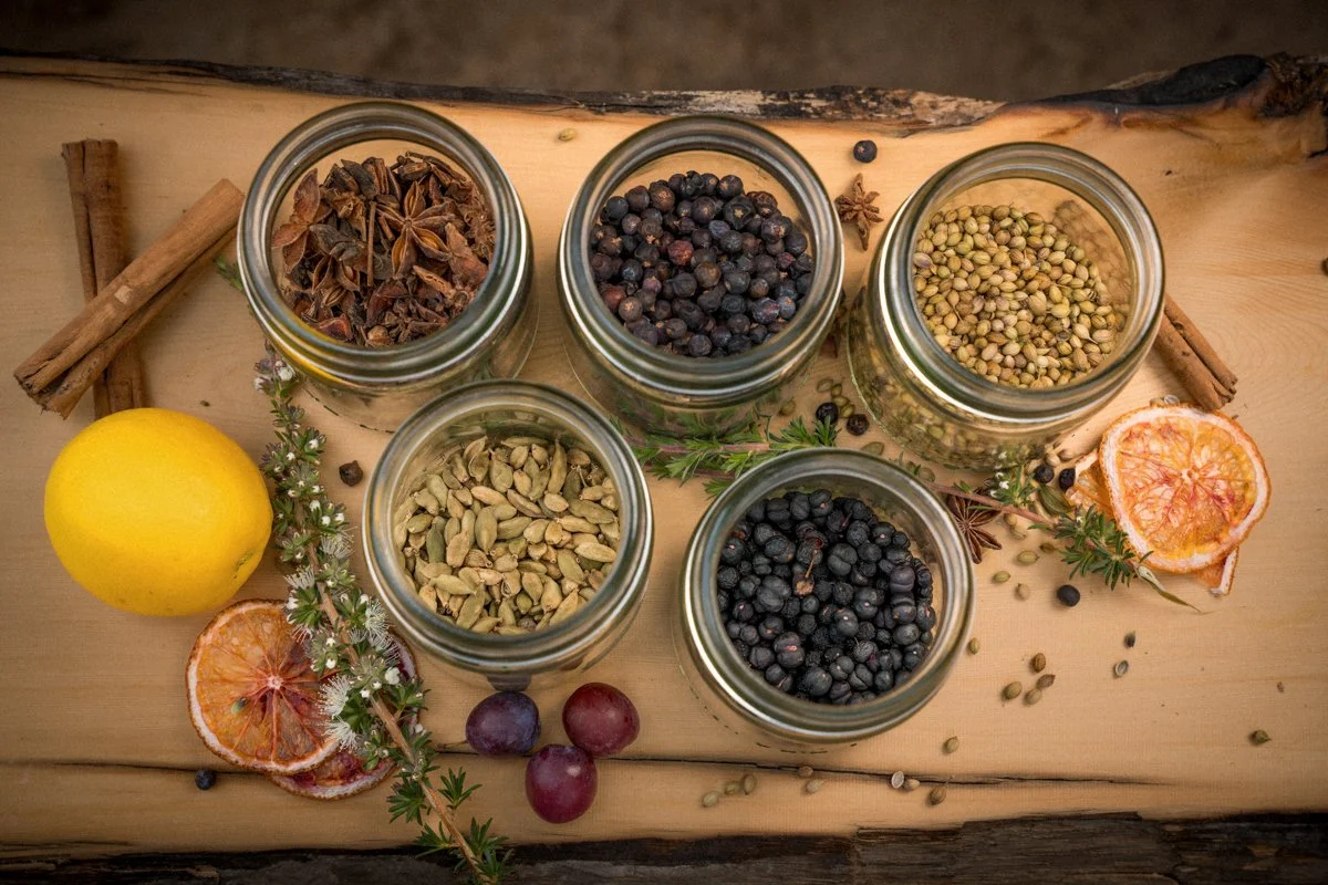 Six glass jars filled with different dried spices and herbs on a wooden surface, surrounded by dried orange slices, grapes, a lemon, cinnamon sticks, star anise, and sprigs of herbs.