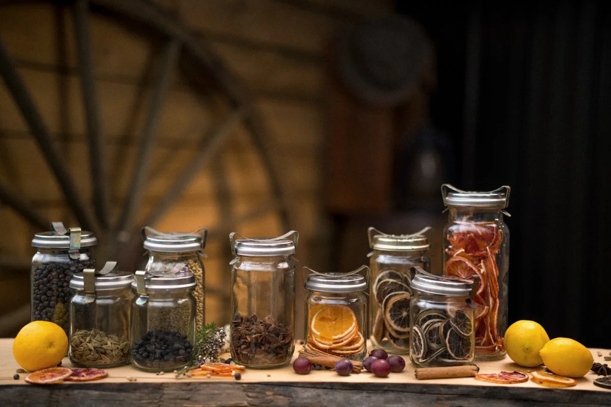 A variety of dried citrus slices, herbs, spices, and berries in glass jars on a wooden surface with lemons and dried fruit pieces scattered around.