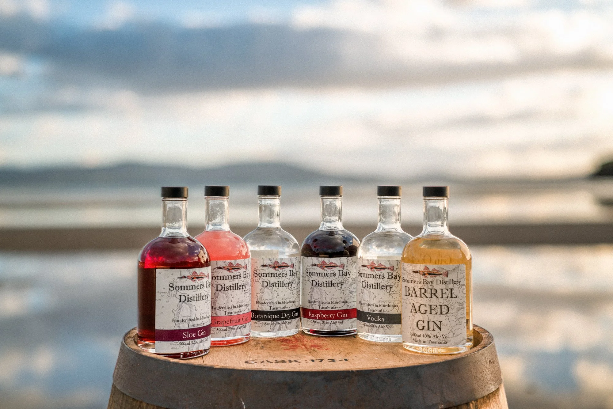 Six bottles of flavored gin from Sommers Bay Distillery are displayed on a wooden barrel with a beach and ocean in the background during sunset or sunrise.
