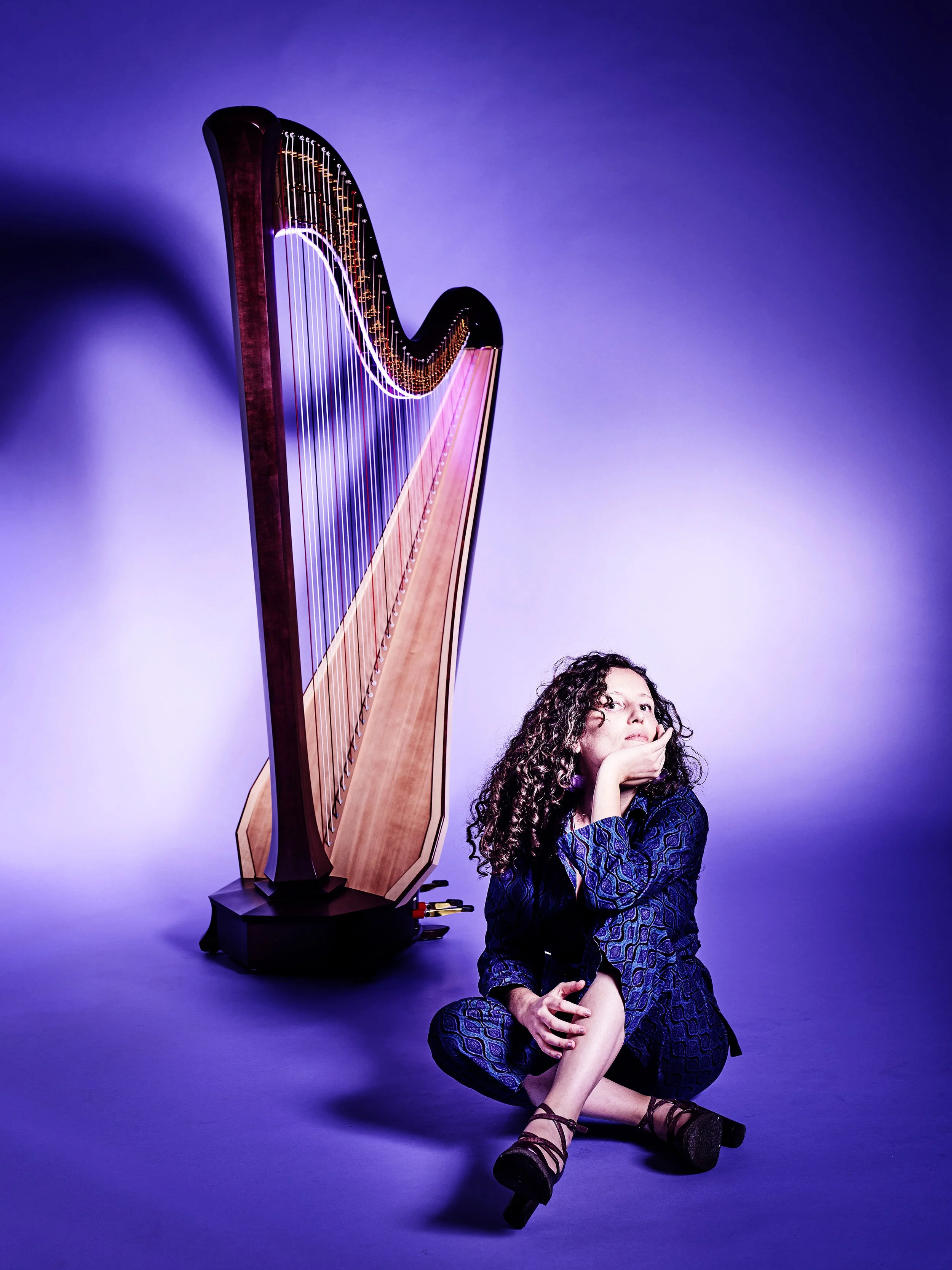Louisic Dulbecco seated on the floor beside a concert harp, purple studio, reflective pose.