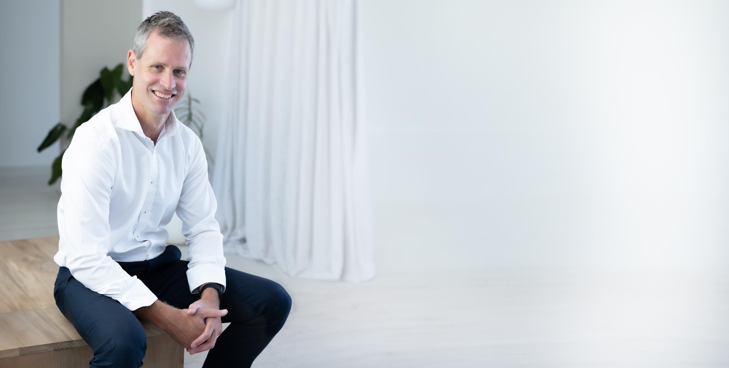 Expert forensic consultant Perth, Dr Clint Hampson, wearing a white collared shirt and black pants, sitting on a wooden table in a bright room with white curtains and a potted plant in the background.