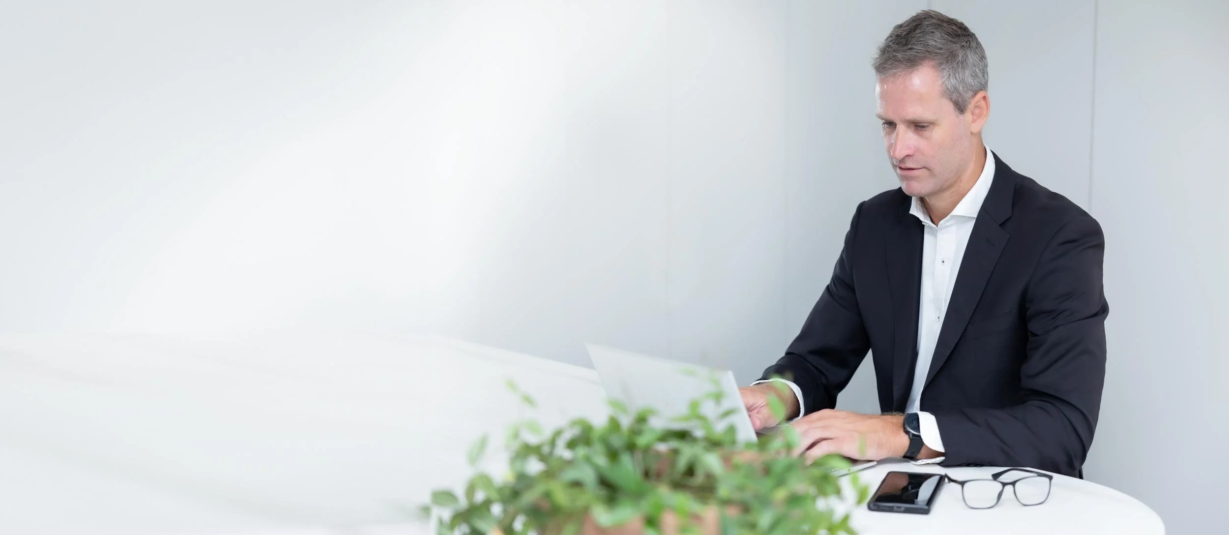 Perth forensic expert, Dr Clint Hampson, in a business suit working on a laptop at a white desk, with a smartphone, glasses, and a plant on the desk.